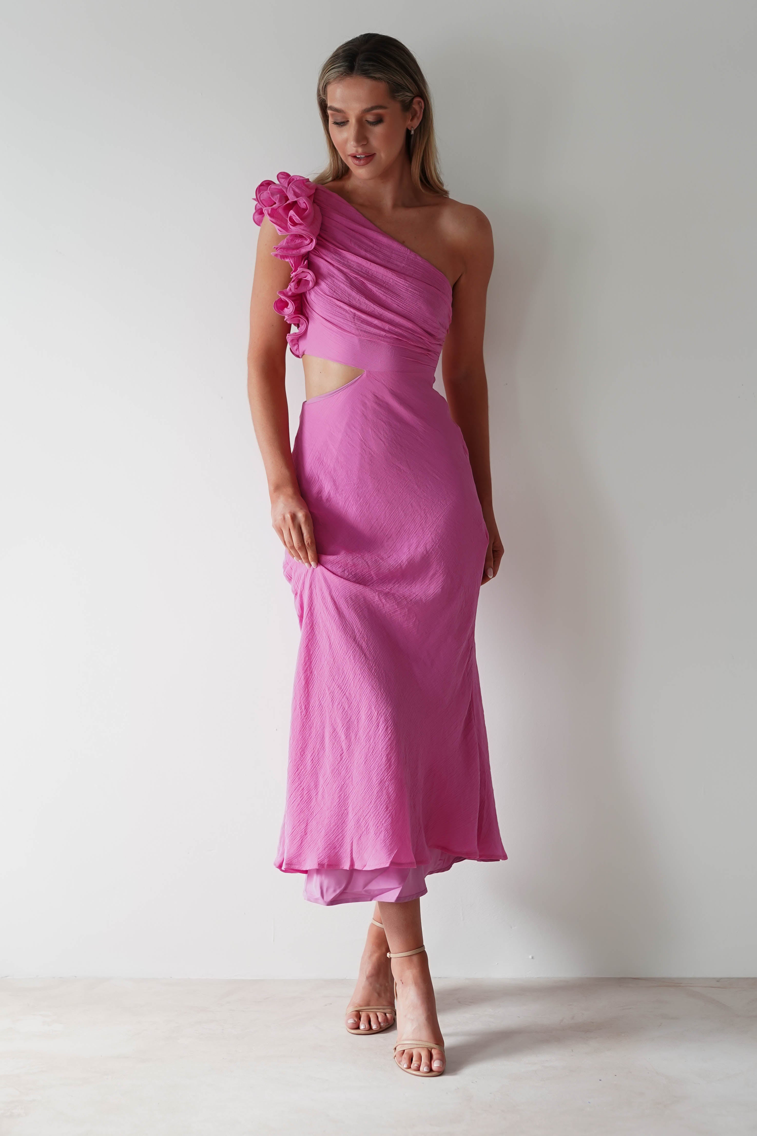 A woman stands against a plain white background in a pink one-shoulder ruffle maxi with floral details on the strap and an elegant midriff cutout. Her long hair cascades down as she wears strappy high heels, looking down with a gentle expression, perfect for special occasions.