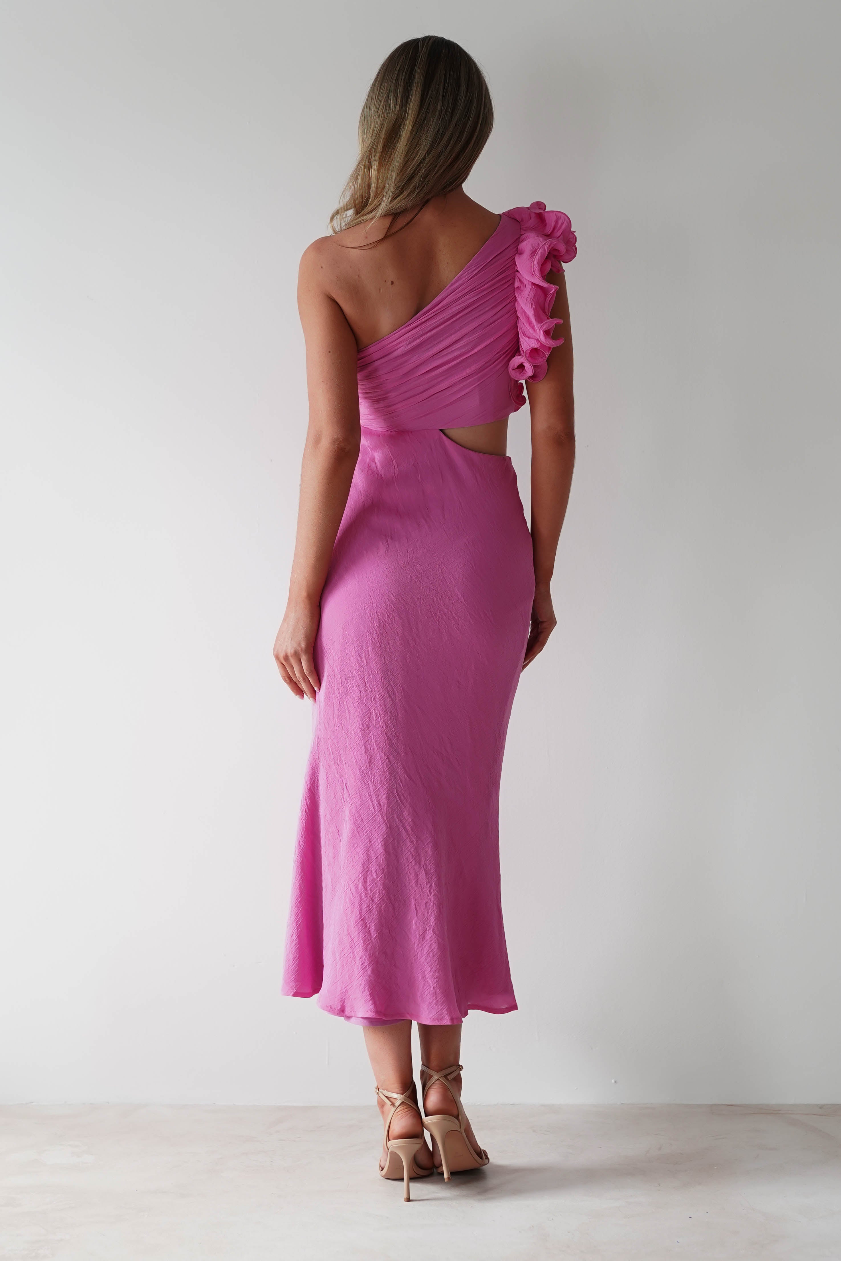 Woman in a pink, one-shoulder ruffle maxi dress with a bare back and midriff cutouts stands facing away in beige heels, perfect for a stylish wedding guest.