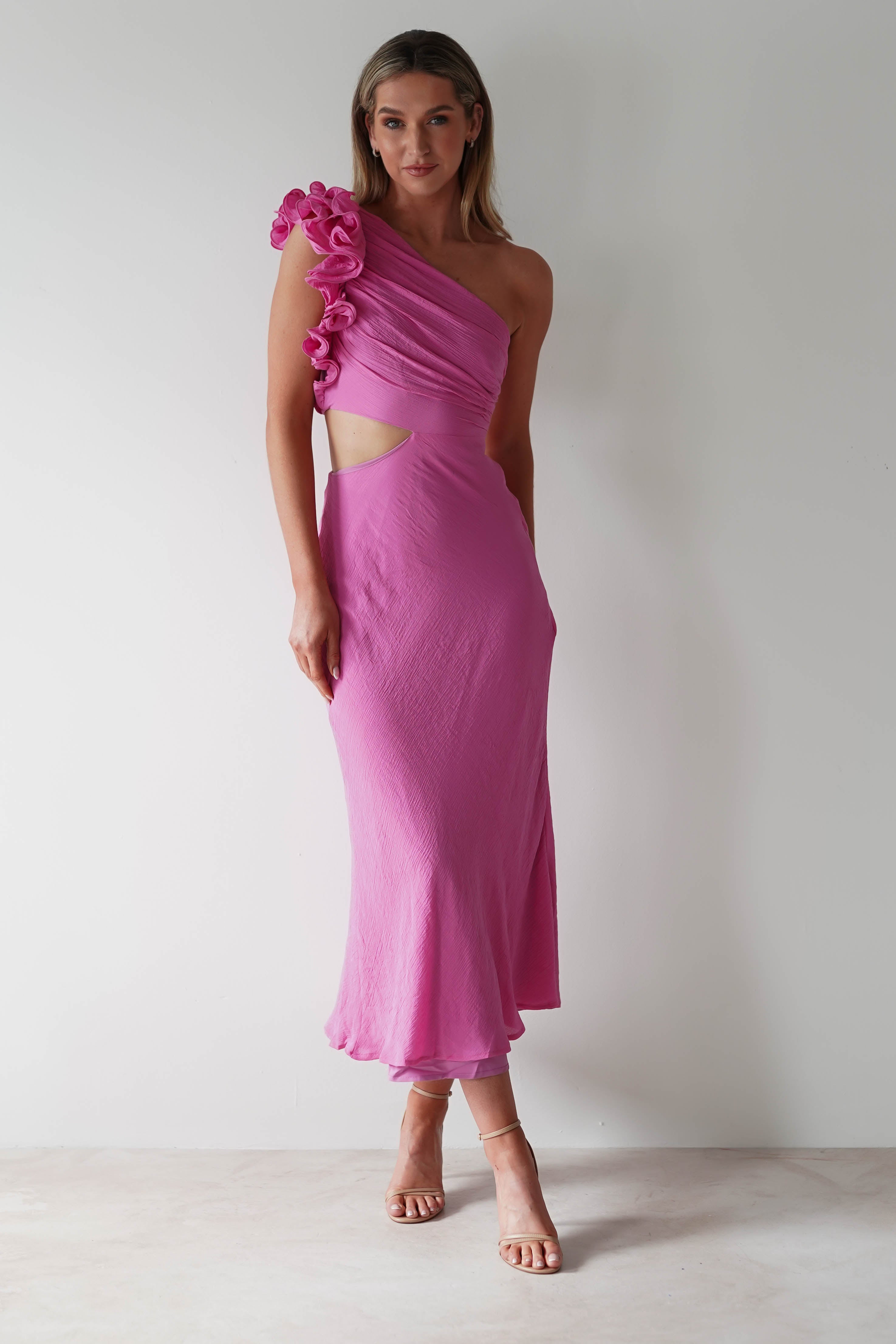 A person wearing a pink one-shoulder ruffle maxi dress with midriff cut-outs stands against a plain white backdrop. They are wearing nude heels and have long hair styled naturally, perfect for a chic wedding guest look.
