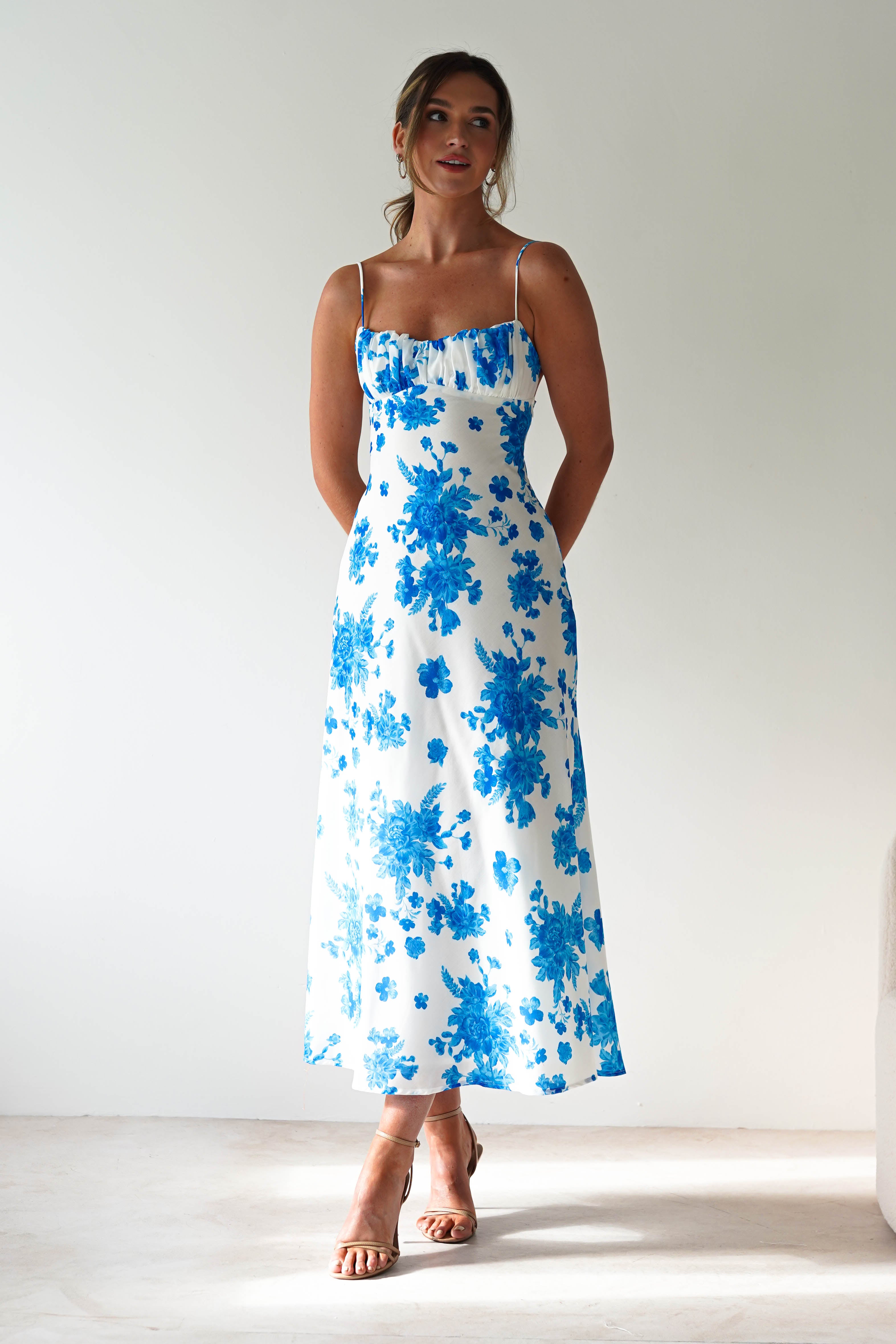 A woman stands indoors wearing a white, blue floral spaghetti strap maxi dress with ruffle detailing on the neckline—perfect for a wedding guest. Her hair is up and she pairs the look with strappy sandals against a plain, neutral background.