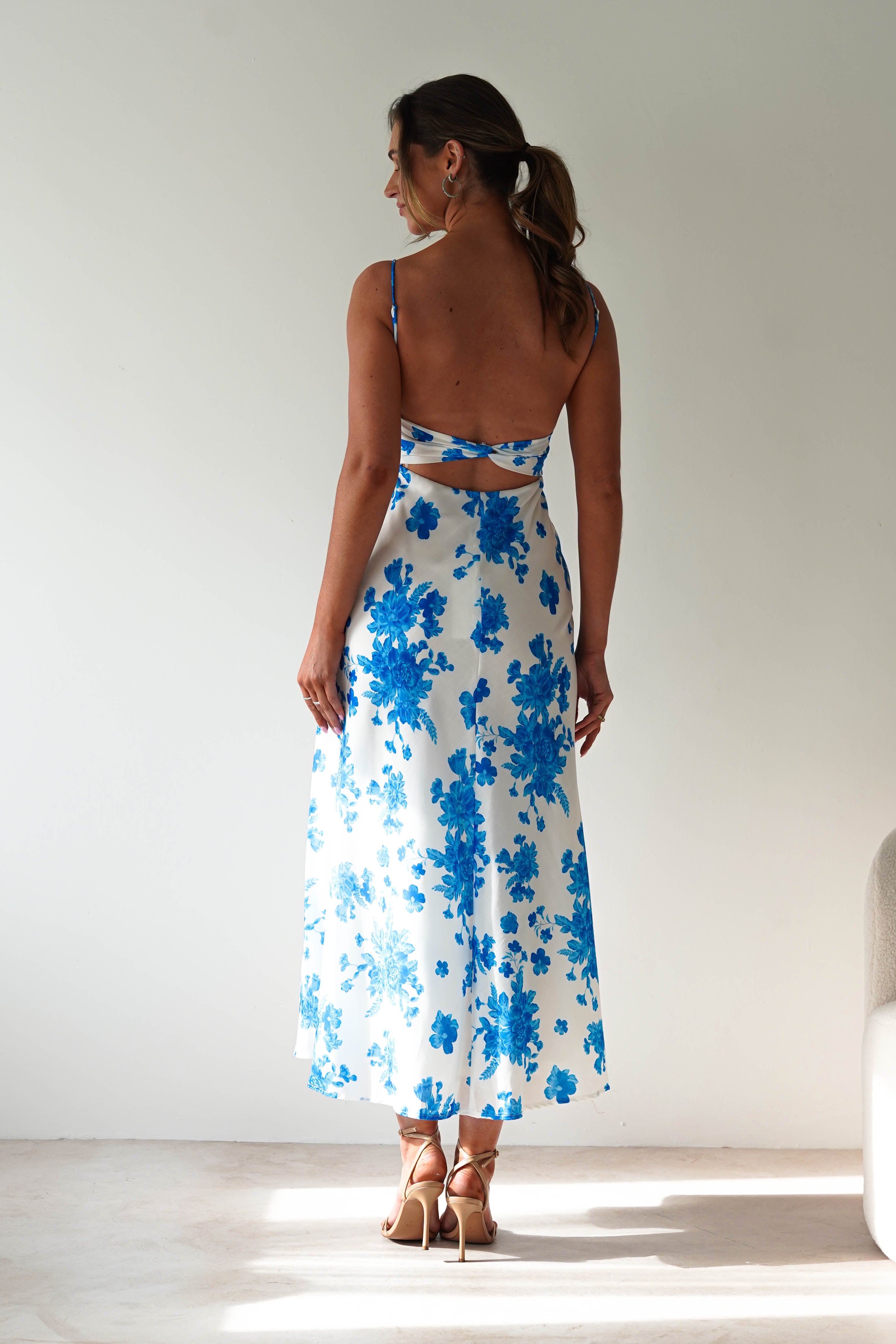 A woman stands indoors facing away, wearing a blue midaxi dress with floral prints, delicate spaghetti straps, and an open back—perfect for a stylish wedding guest. Her brown hair is in a ponytail with a white ribbon, and she wears beige heels.