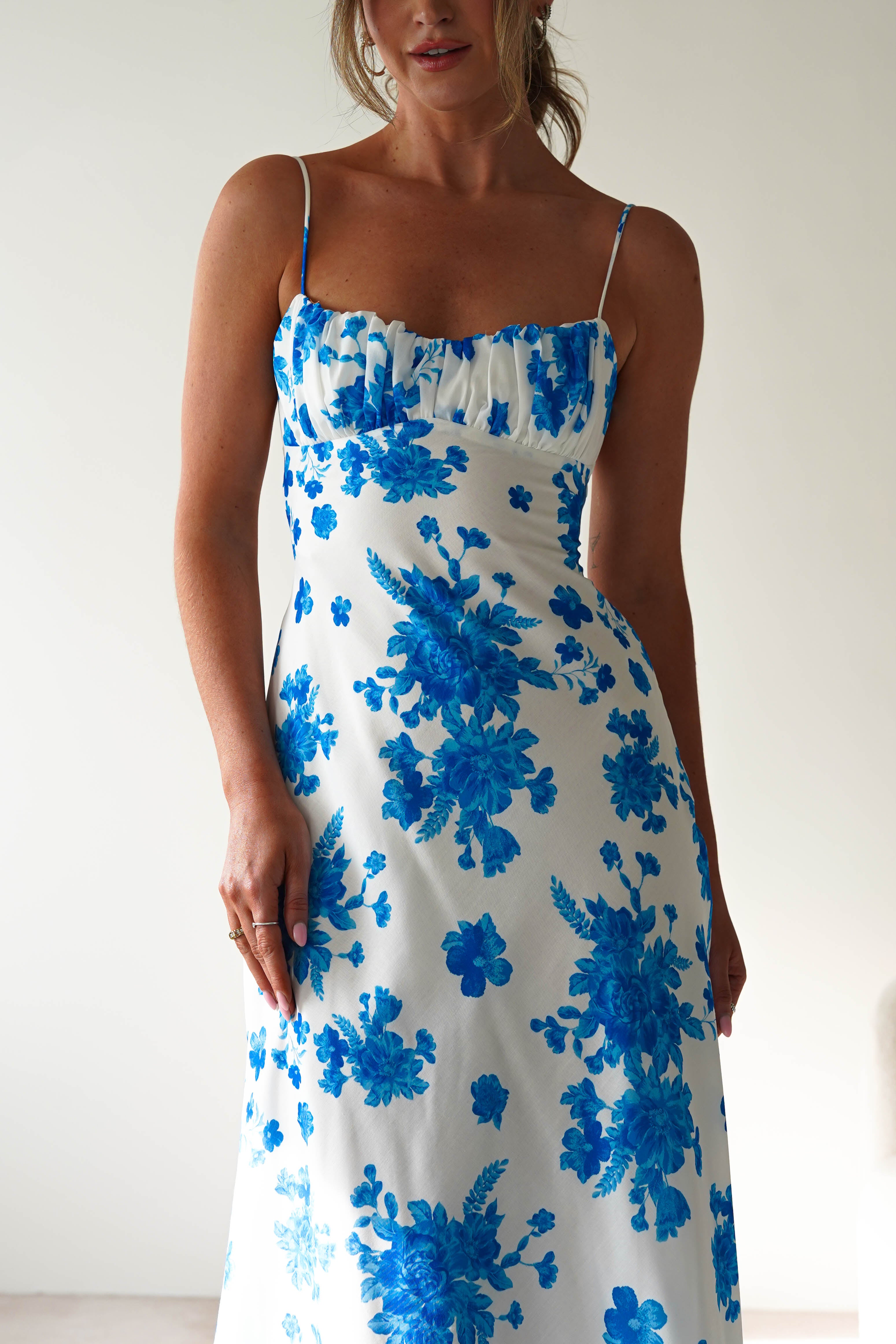A woman wearing a white spaghetti strap dress with a blue floral pattern stands against a plain light-colored background. The blue midaxi dress features a fitted bodice and a flowing skirt, perfect for a wedding guest look.