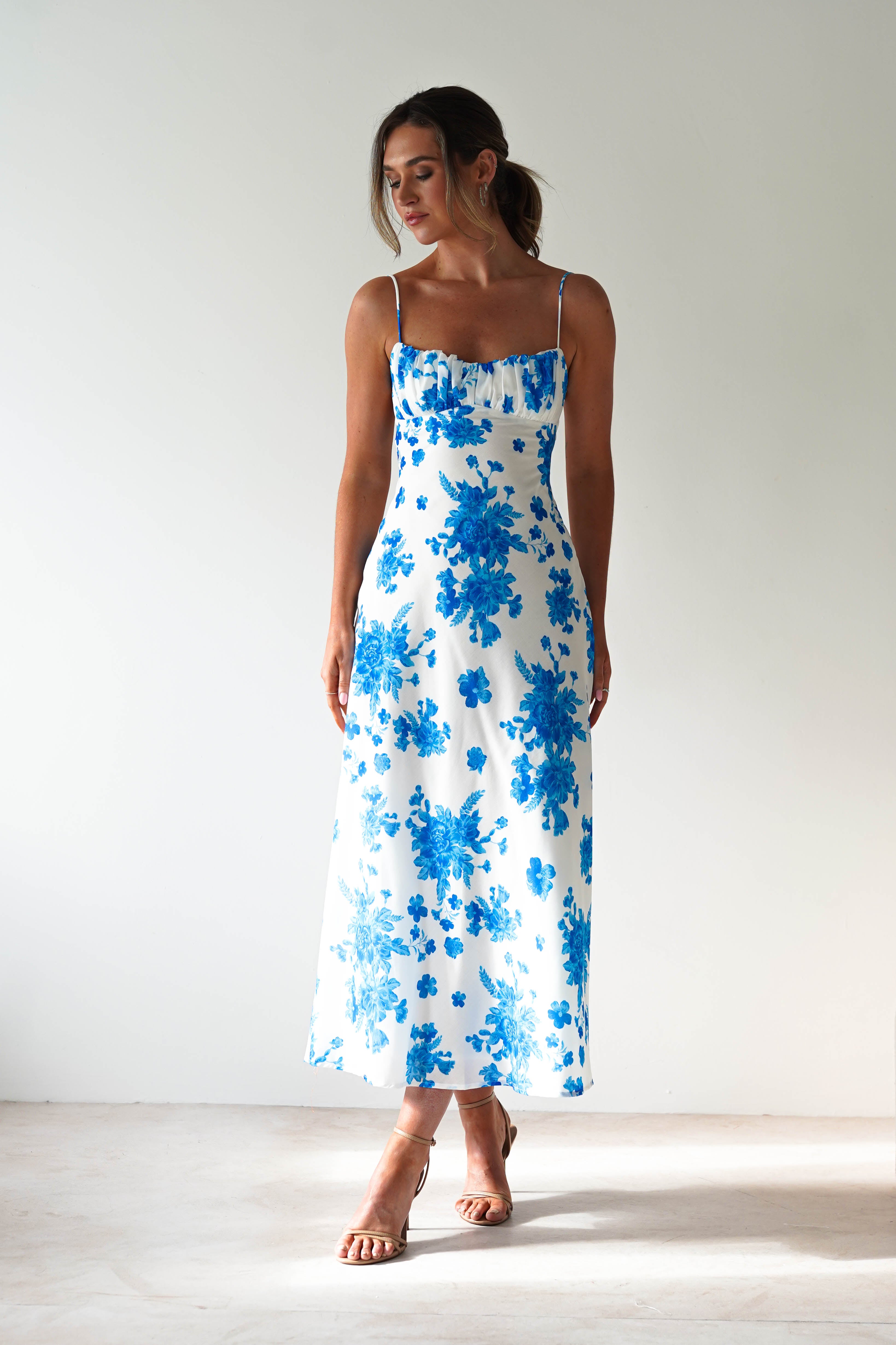 A woman stands against a plain light background in a blue midaxi sleeveless dress with blue floral patterns and delicate spaghetti straps. Her hair is tied back, and she completes the wedding guest look with strappy beige heels.