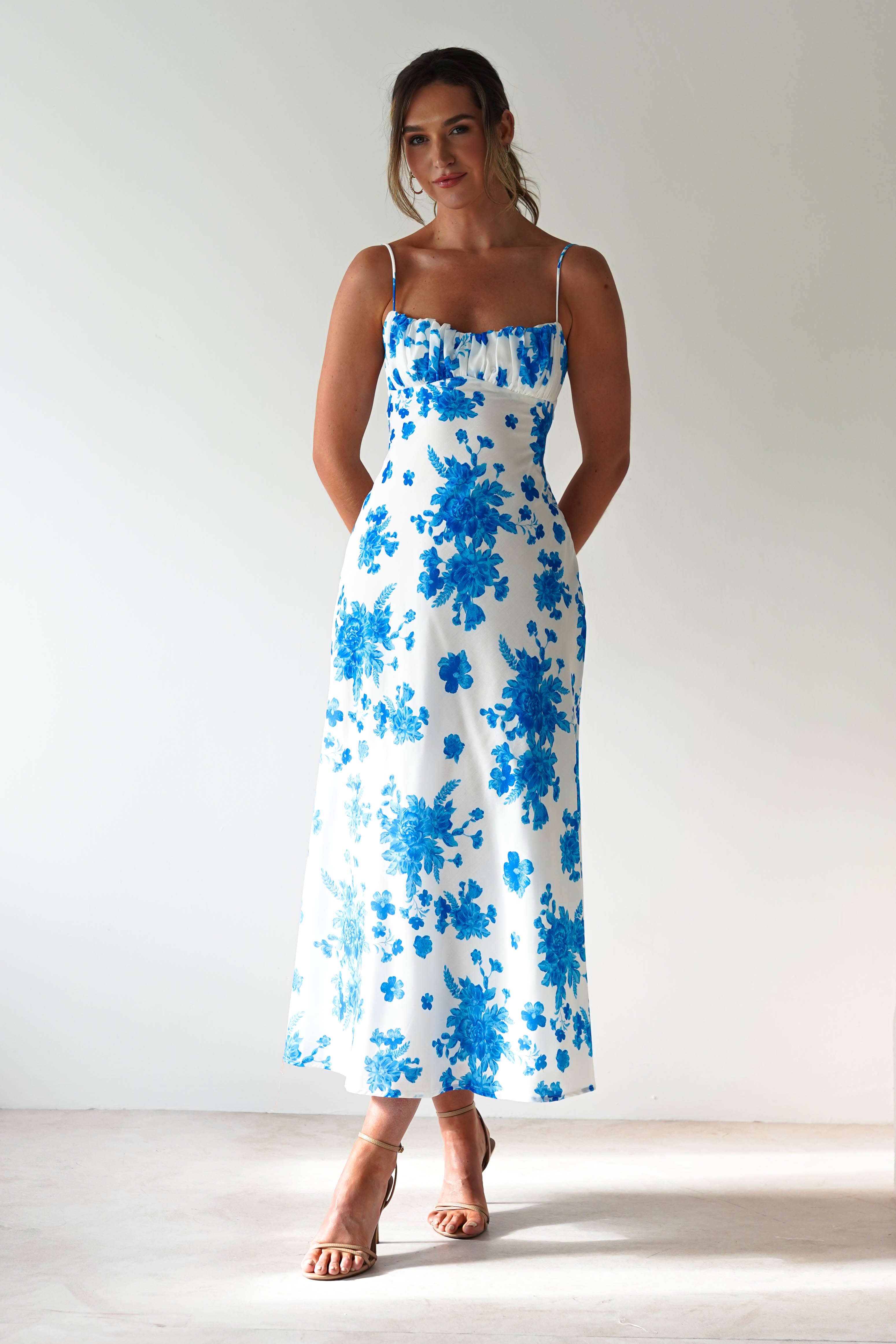 A woman stands against a plain light background in a blue floral spaghetti strap dress, perfect for a wedding guest. She pairs the white midaxi with nude heels, her hair tied back and hands behind her back.