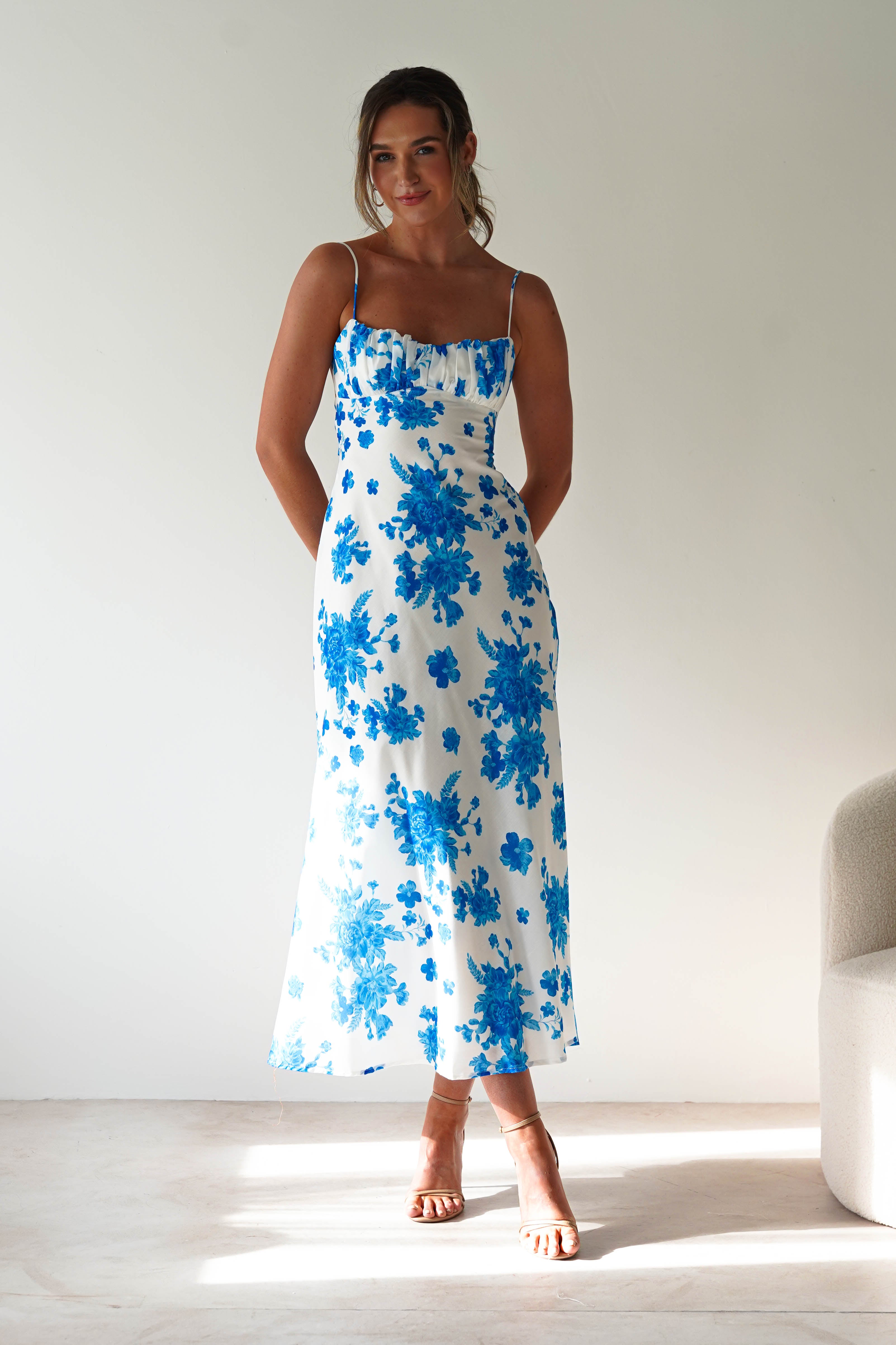 A woman stands indoors in a blue midaxi dress with spaghetti straps and white floral patterns. She poses with her hands in her pockets as natural light shines on her from the left, while a light-colored chair is partially visible on the right.