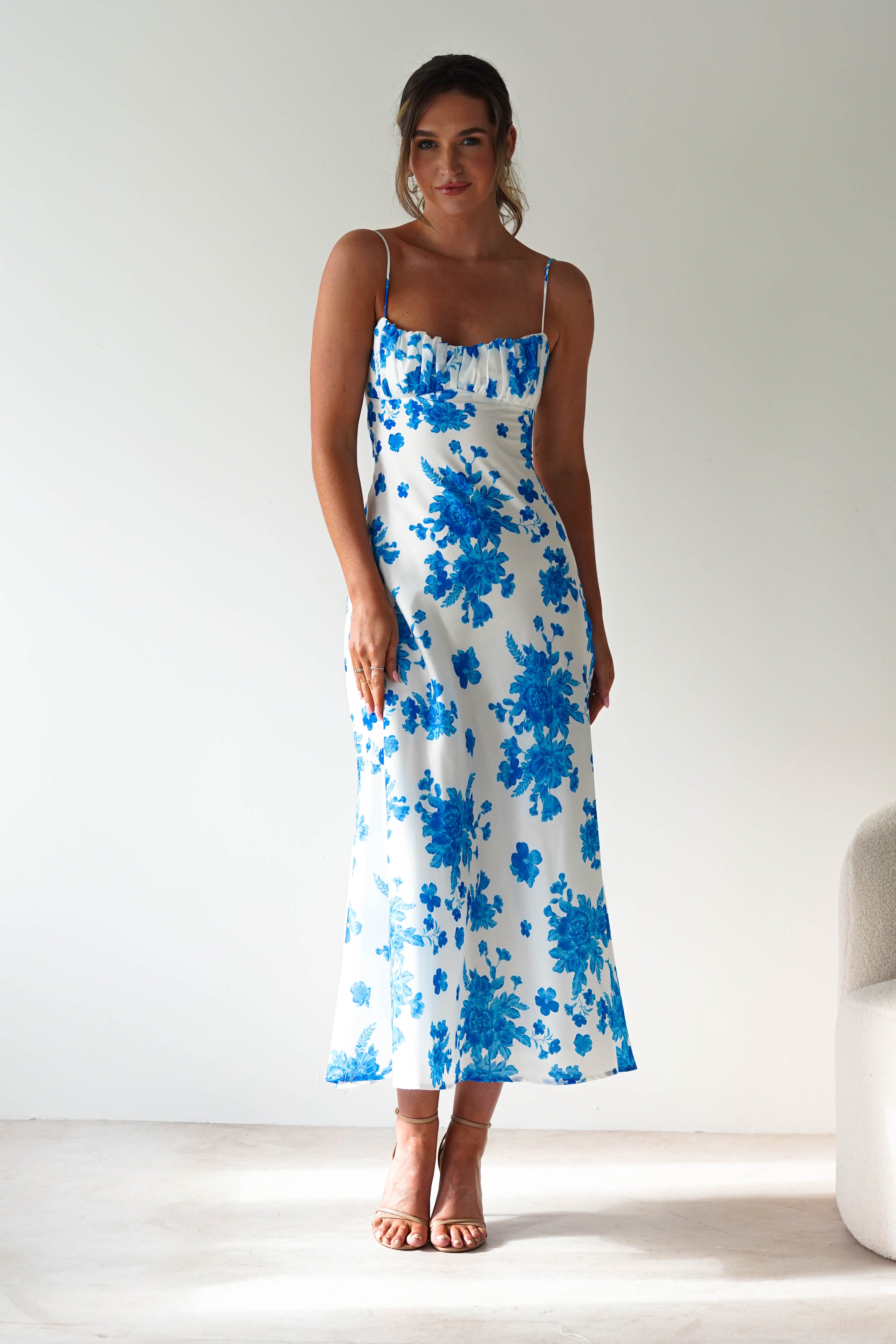 A woman stands indoors against a plain light-colored wall, wearing a blue midaxi spaghetti strap dress with bright floral patterns. Her hair is tied back and she completes this perfect wedding guest look with nude heels.