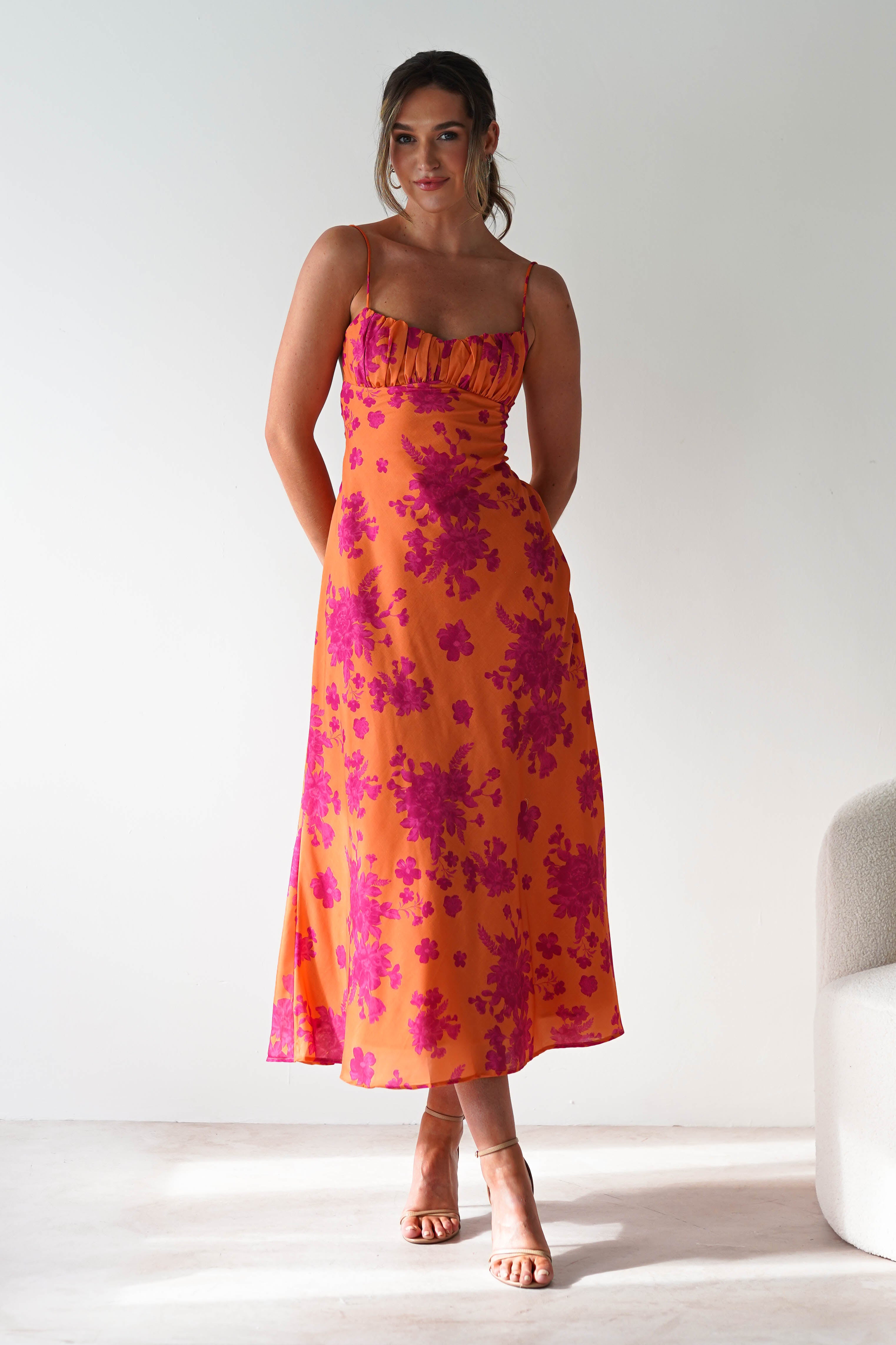 A woman stands indoors wearing a vibrant spaghetti strap orange dress with pink floral patterns, paired with nude heels. She smiles with her hands behind her back, next to a light-colored chair.