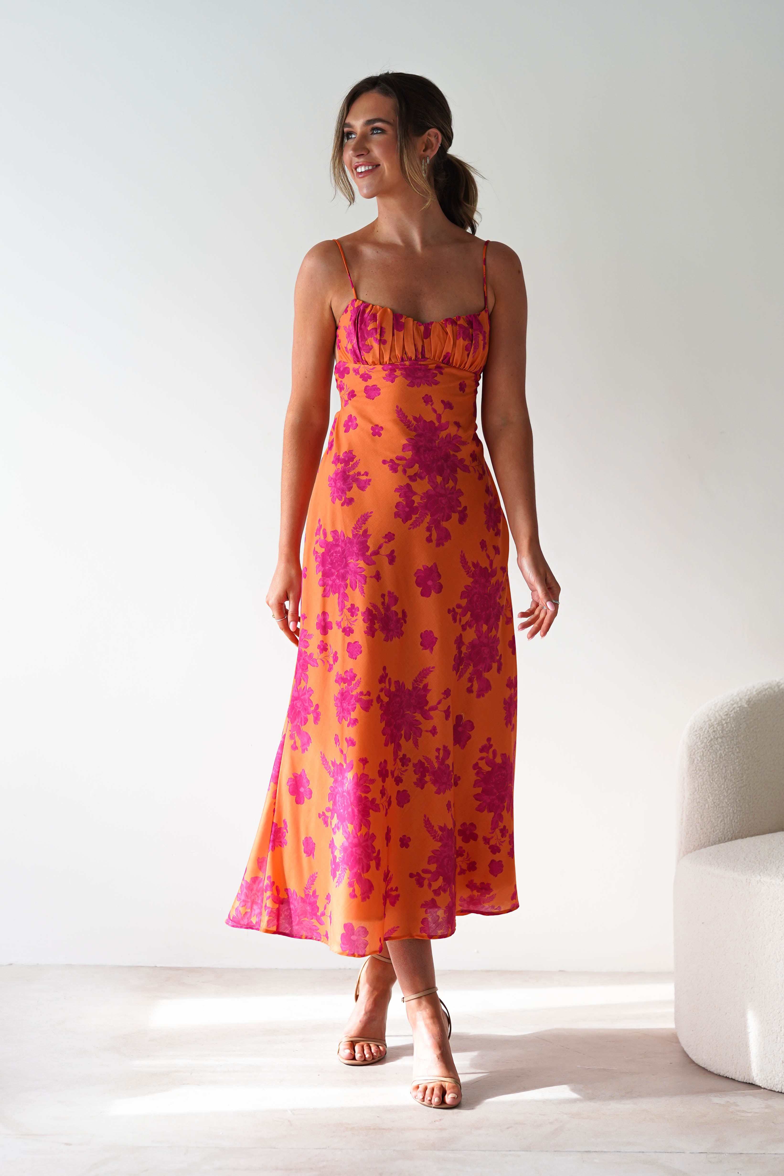 A woman stands smiling in a bright room, wearing a vibrant wedding guest dress with pink floral patterns and spaghetti straps. She has her hair tied back and is positioned near a white, curved chair.
