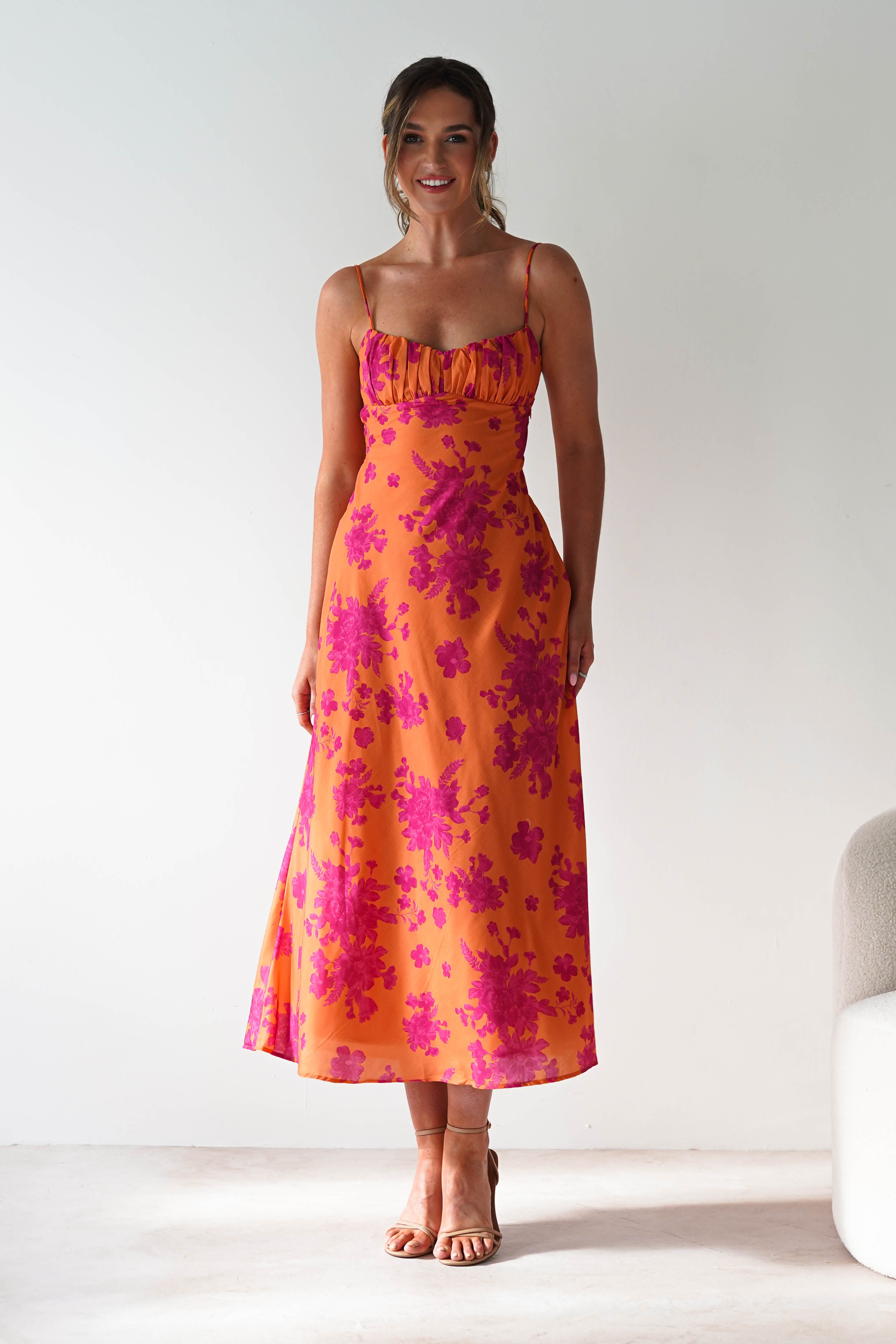 A woman wearing a spaghetti strap, orange dress with pink floral patterns stands indoors on a light floor, smiling, with her hair tied back and hands relaxed at her sides.