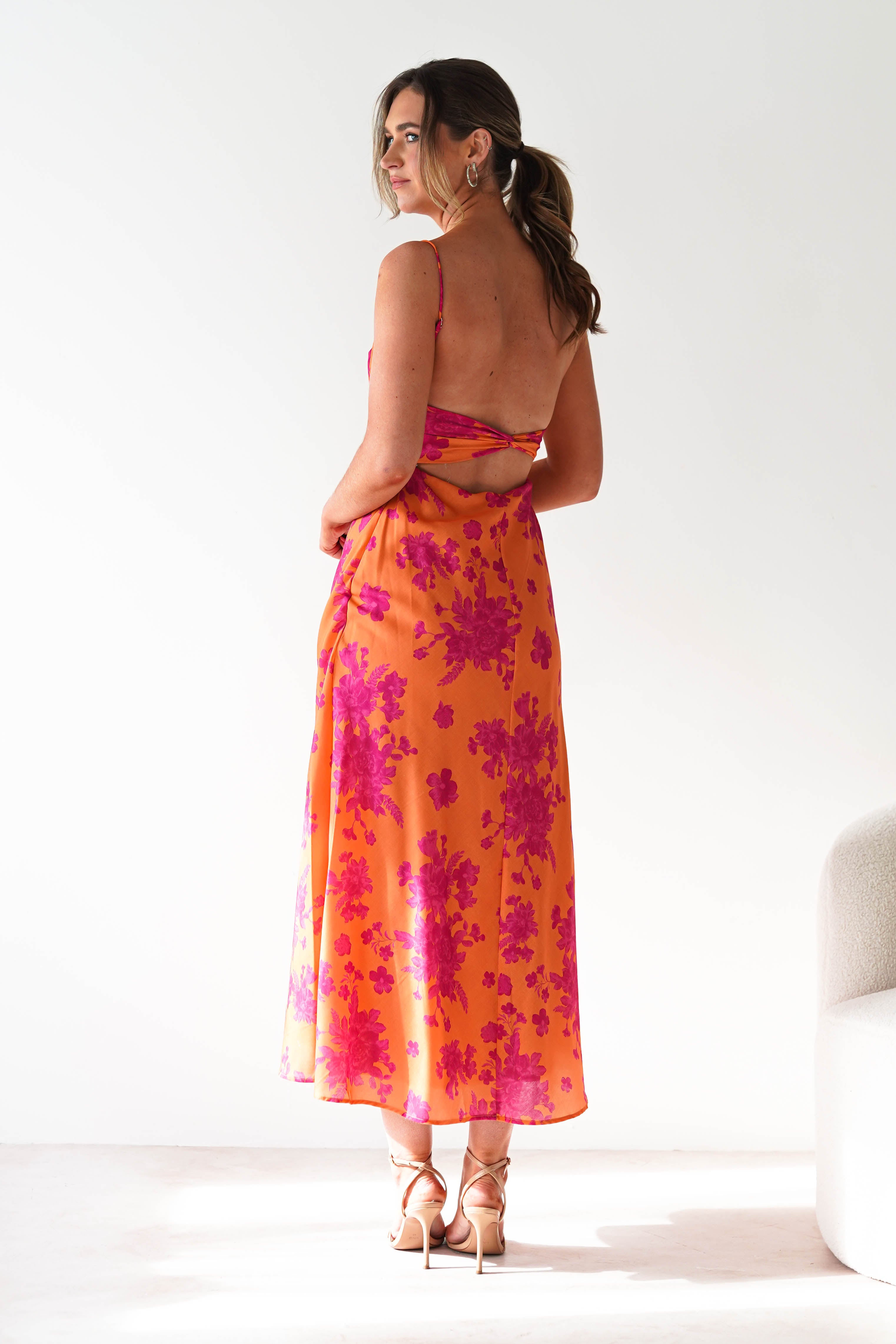 A woman stands sideways, looking over her shoulder, wearing a sleeveless orange wedding guest dress with pink floral patterns and strappy heels. Her hair is tied back and she is in a brightly lit, minimalist room.
