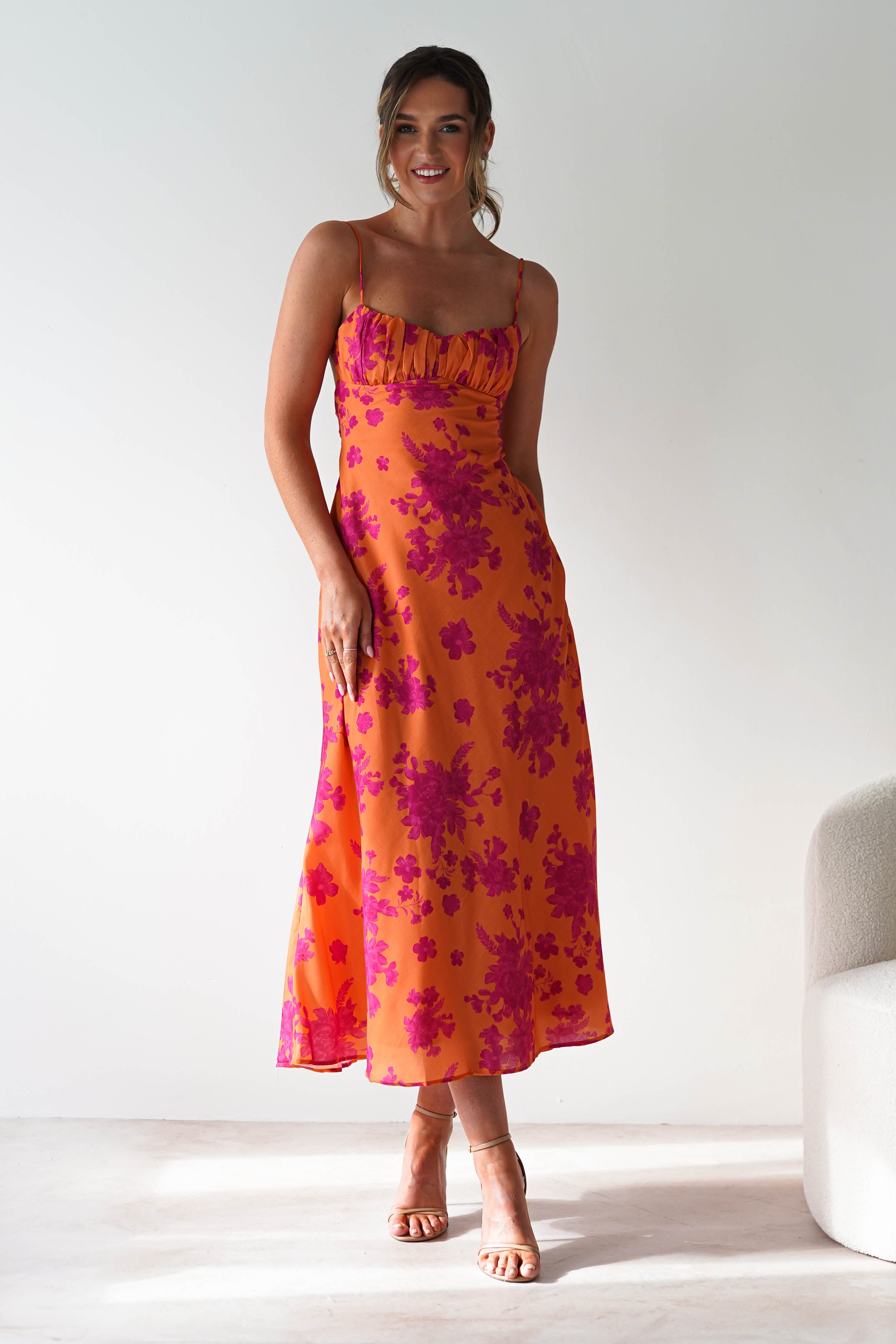A woman stands smiling in a sleeveless orange dress with pink floral patterns, thin spaghetti straps, and a fitted bodice—perfect as a wedding guest dress. She wears nude heels next to a light-colored modern chair against a plain white background.