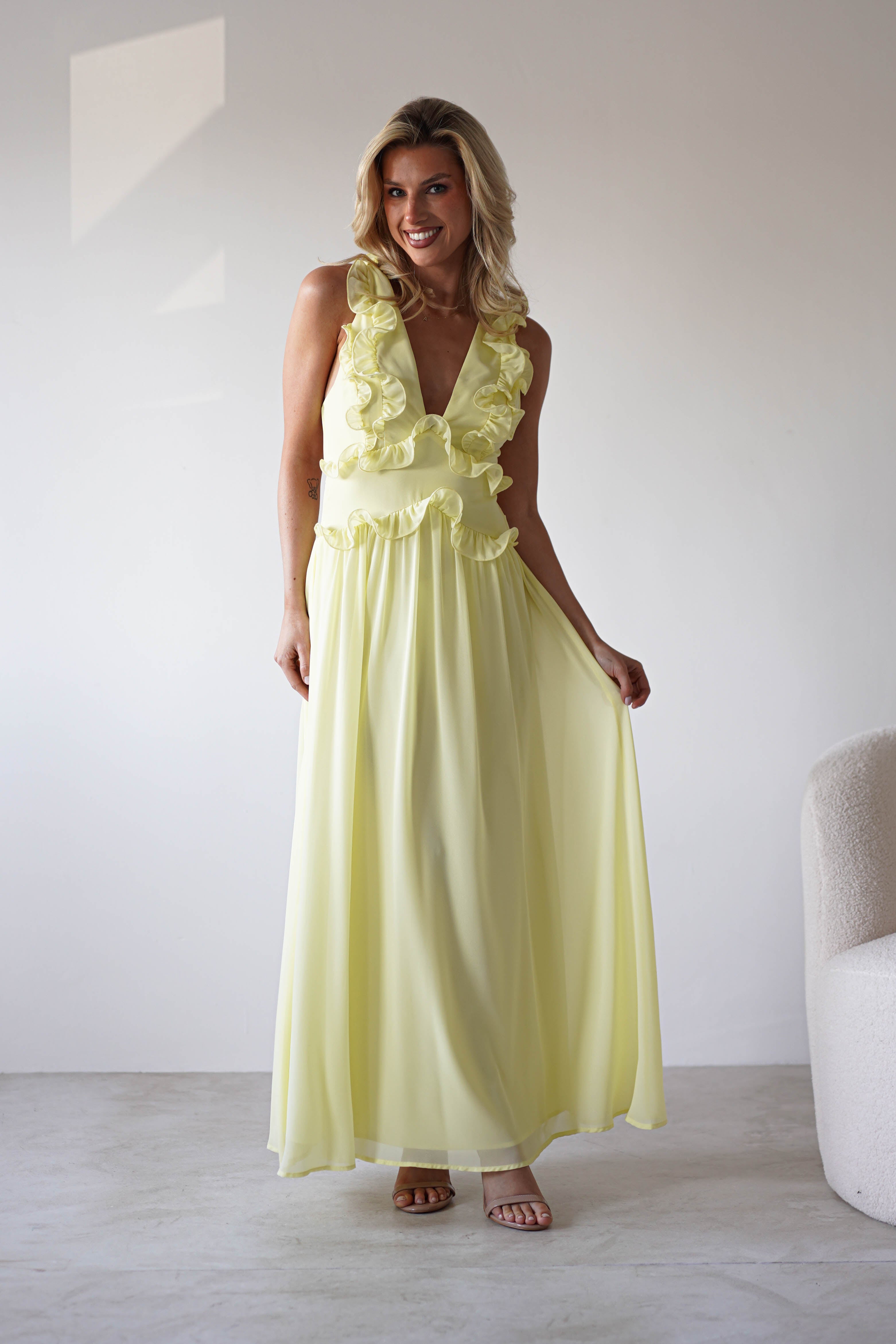 A woman in a light yellow chiffon maxi dress with ruffle straps stands indoors, smiling as she gently holds out her skirt. The simple background with white walls and a beige chair highlights her look, perfect for special occasions.