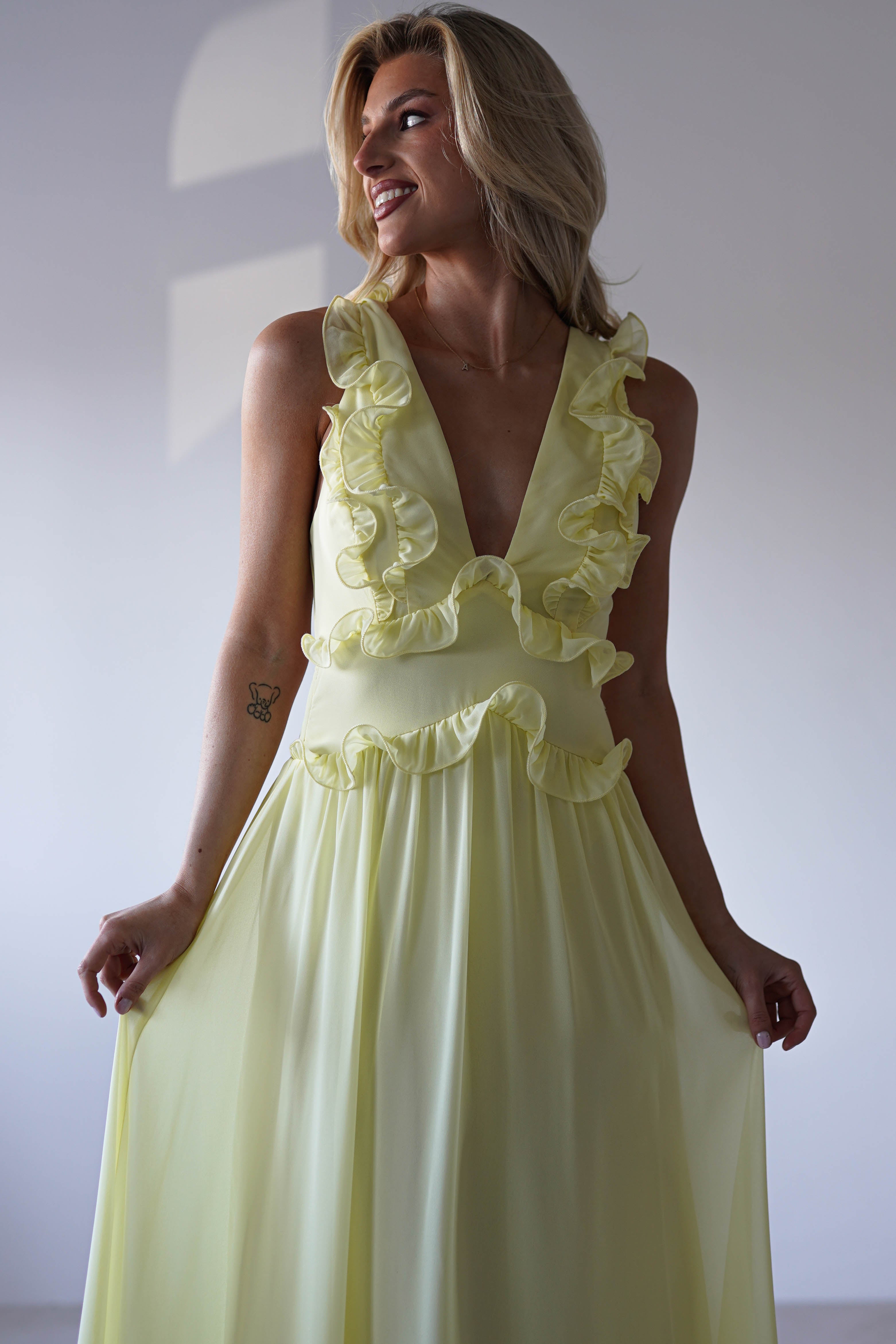 A woman in a sleeveless, pale yellow chiffon maxi dress with ruffle straps stands smiling, holding the sides of her skirt. She has blonde hair and a small tattoo on her left arm, softly lit against a plain background—perfect for special occasions.
