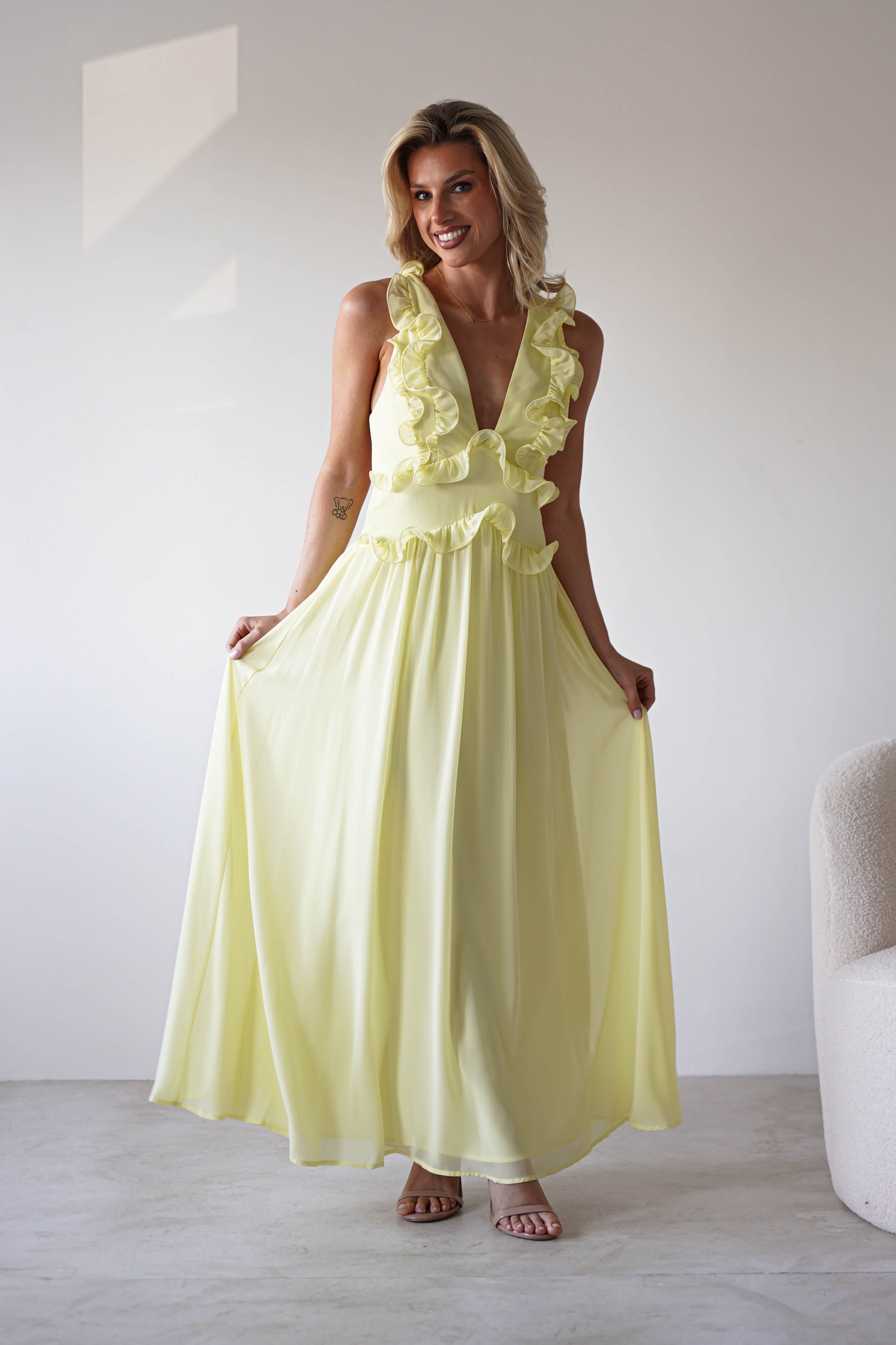 A woman stands indoors wearing a pale yellow chiffon maxi dress with ruffle straps and a deep V-neckline, smiling as she lightly holds the sides of her skirt—a perfect look for special occasions. The background is minimal, with a white wall and light chair.