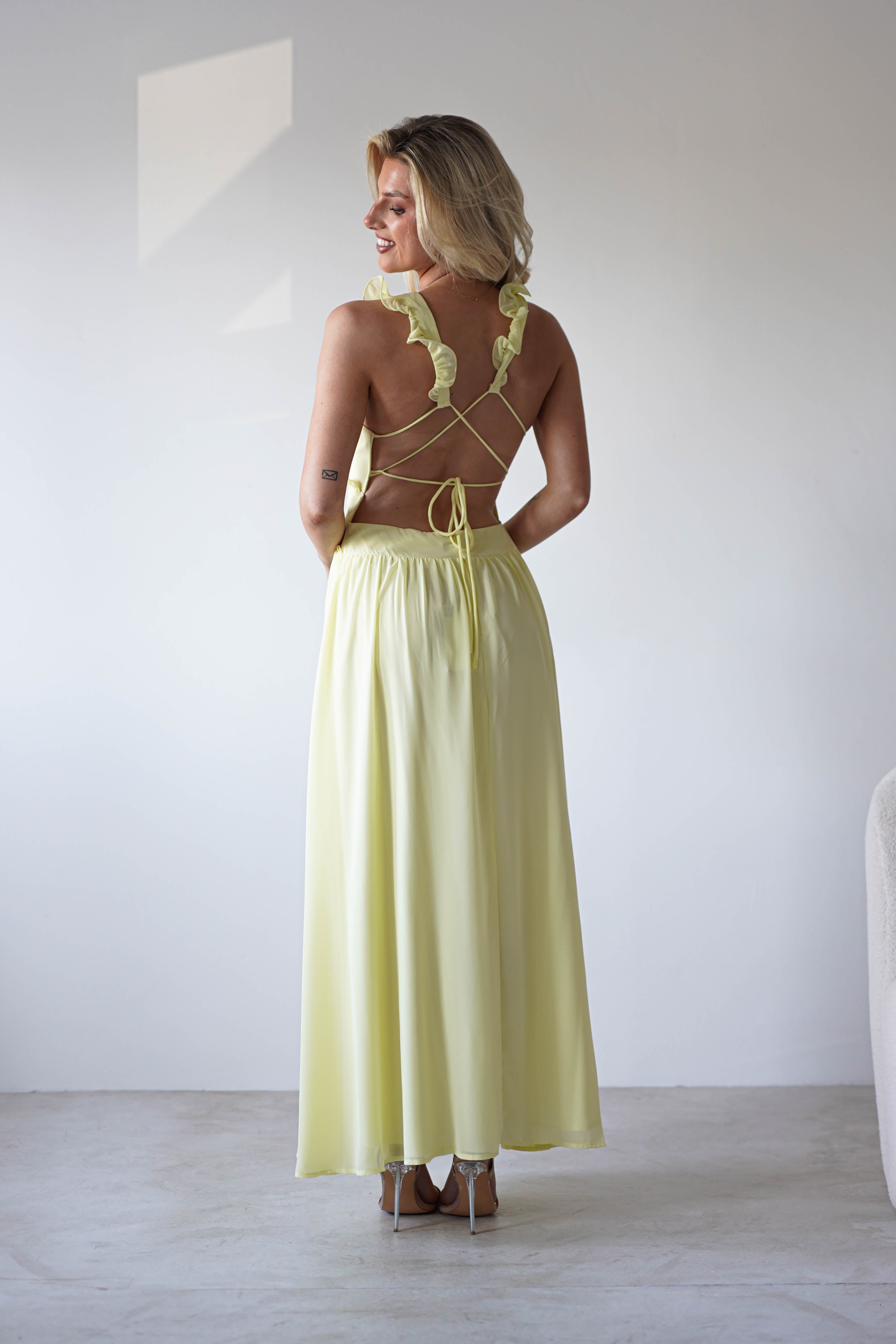 A woman wearing a long, light yellow chiffon maxi dress with an open back and ruffle straps stands indoors on a light floor, facing away from the camera and smiling over her shoulder—perfect for special occasions.