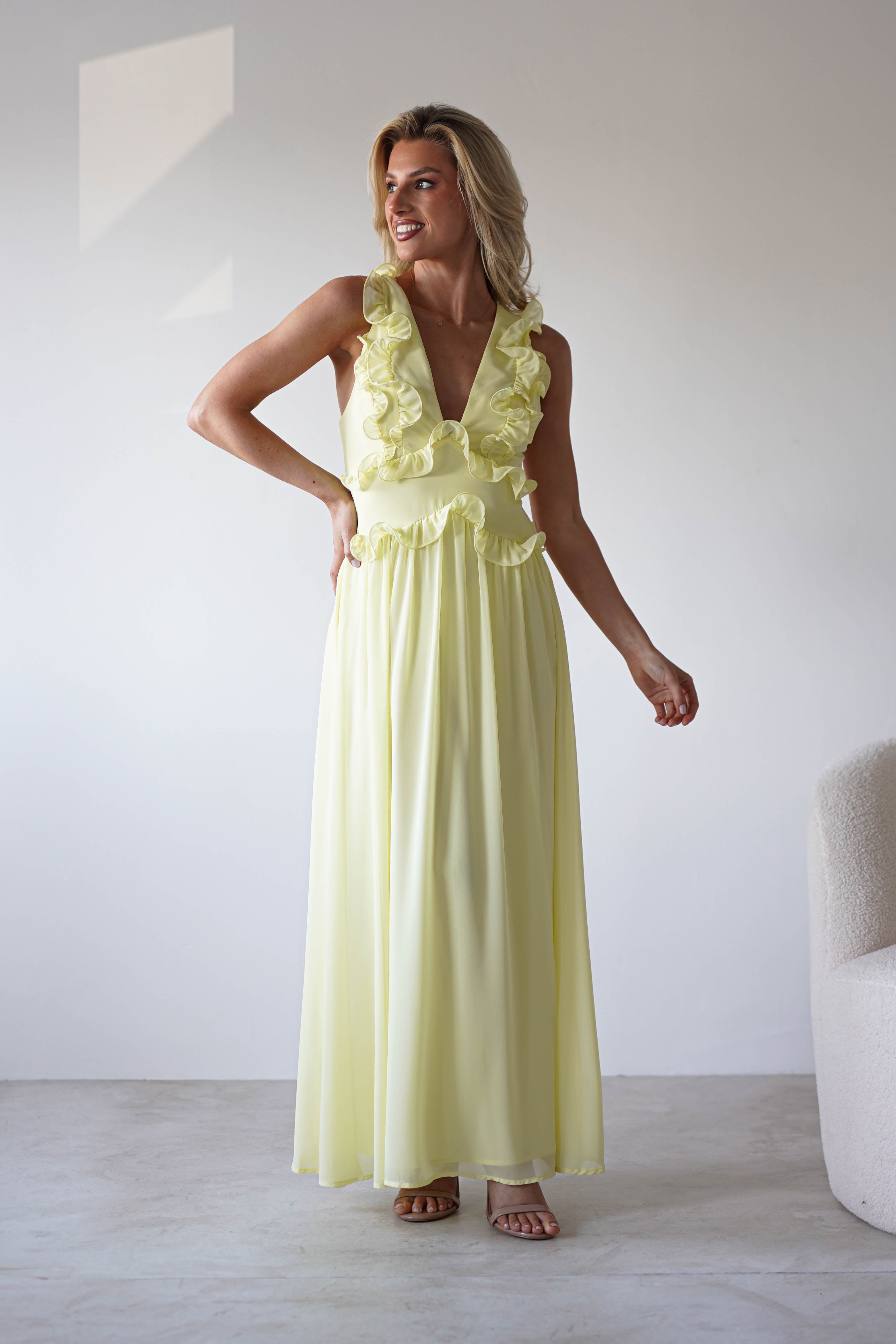 A woman wearing a sleeveless chiffon maxi dress with ruffle details stands indoors beside a white textured chair, smiling and looking to her left—perfect for special occasions.