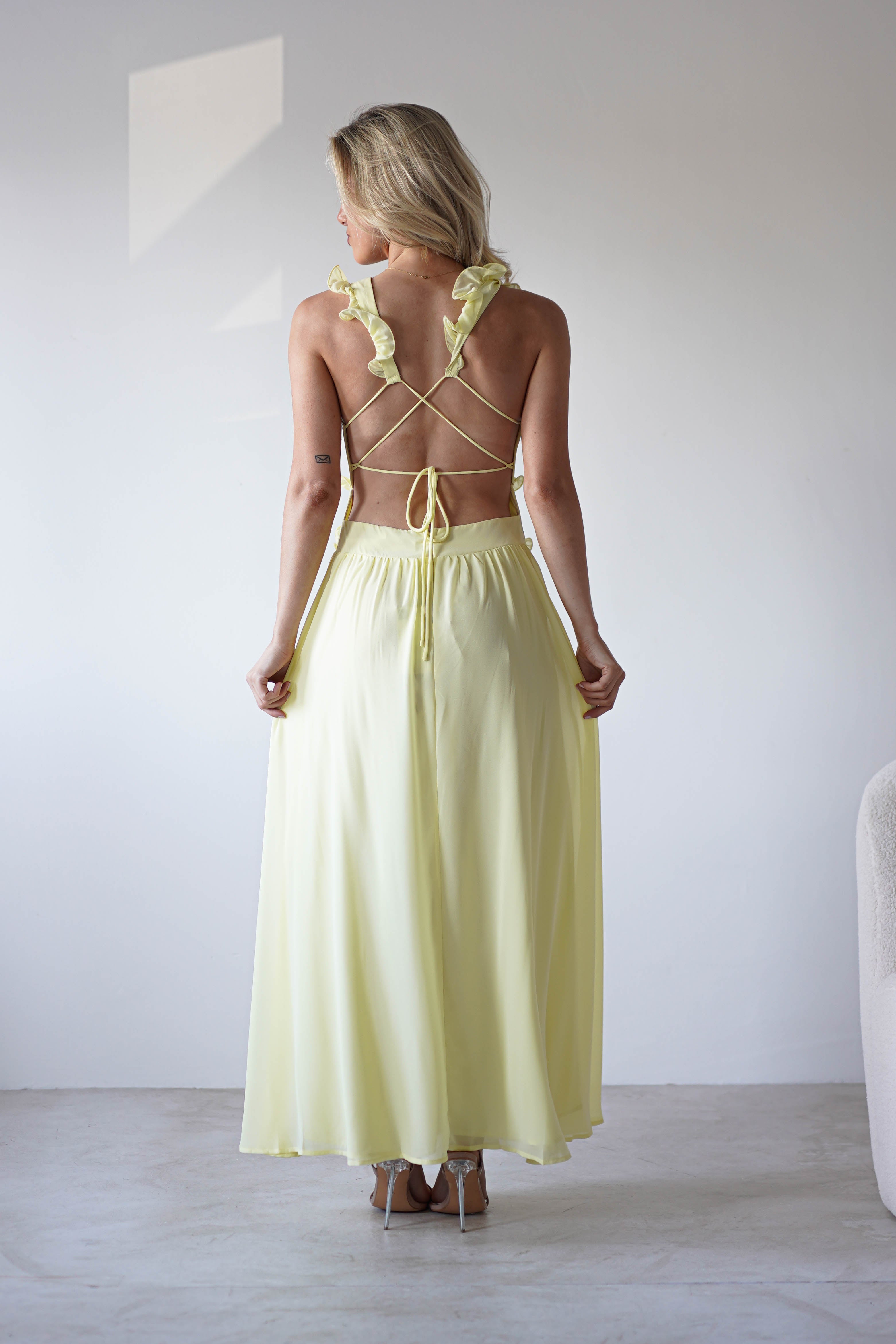 A woman stands indoors facing away, wearing a light yellow chiffon maxi dress with ruffle straps and crisscrossed back, holding the sides of her skirt. The softly lit, minimal background highlights this look for special occasions.