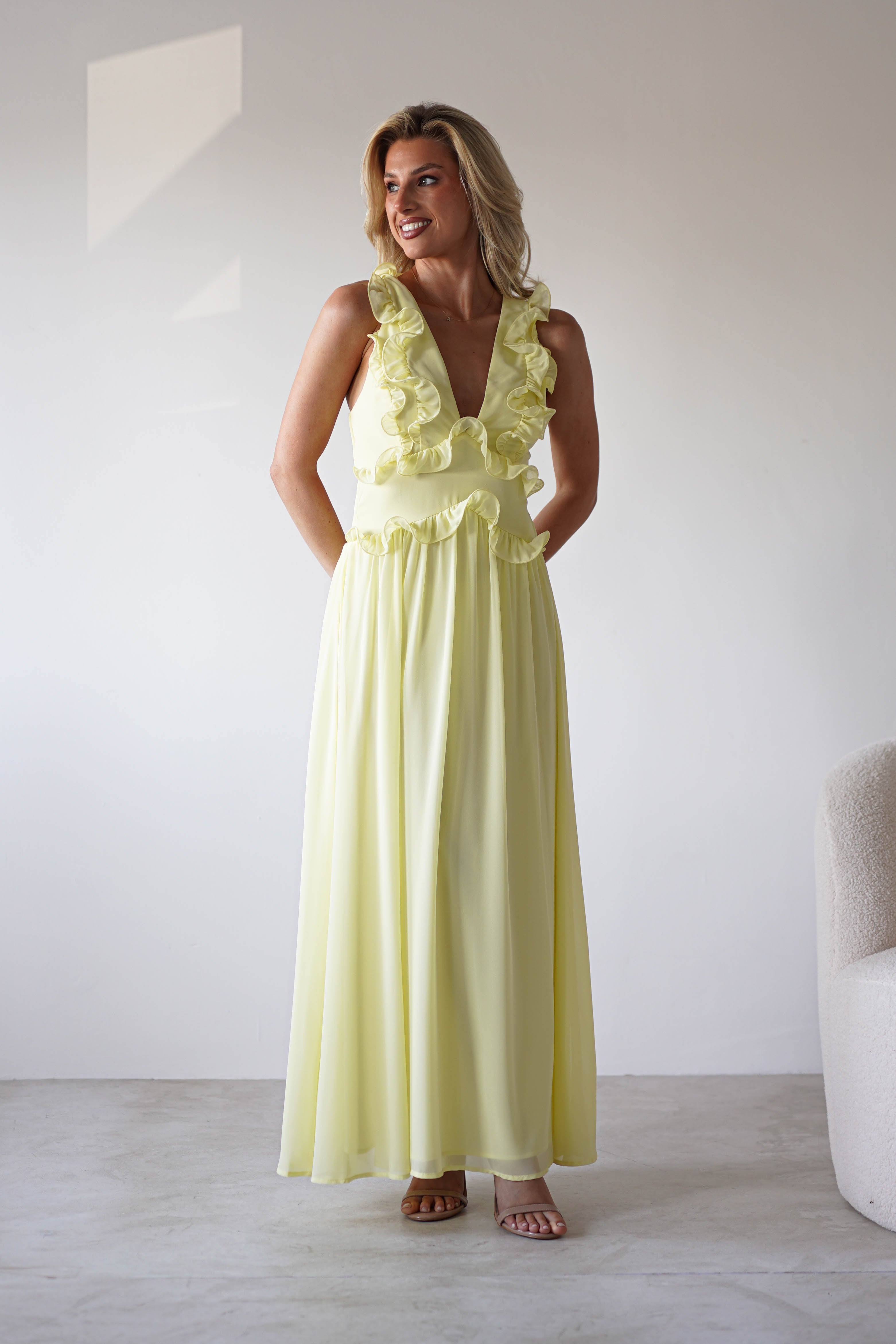 A woman in a light yellow chiffon maxi dress with ruffle straps stands smiling with her hands behind her back in a bright, minimalist room—perfect for special occasions.