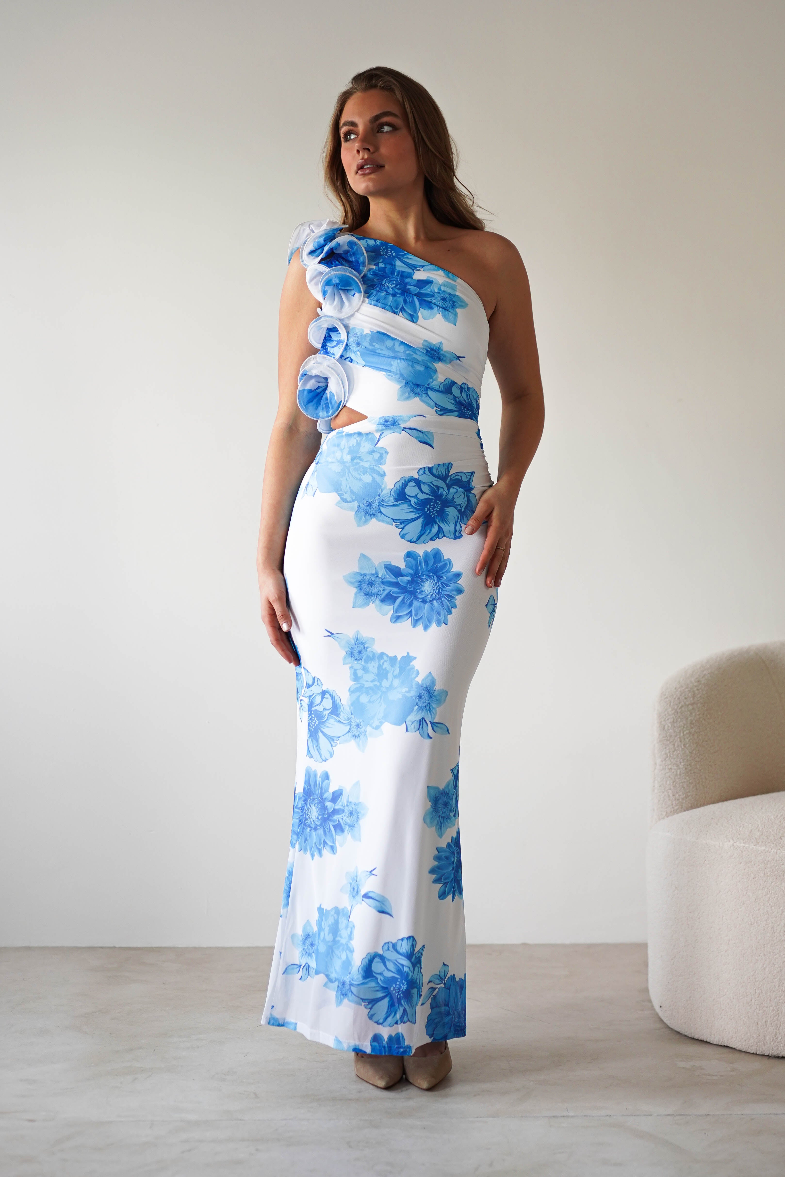 A woman stands in a white room wearing a long, fitted wedding guest dress with a summer blue print, featuring a one-shoulder design and decorative fabric rosettes. She has long brown hair and is standing near a curved white chair.