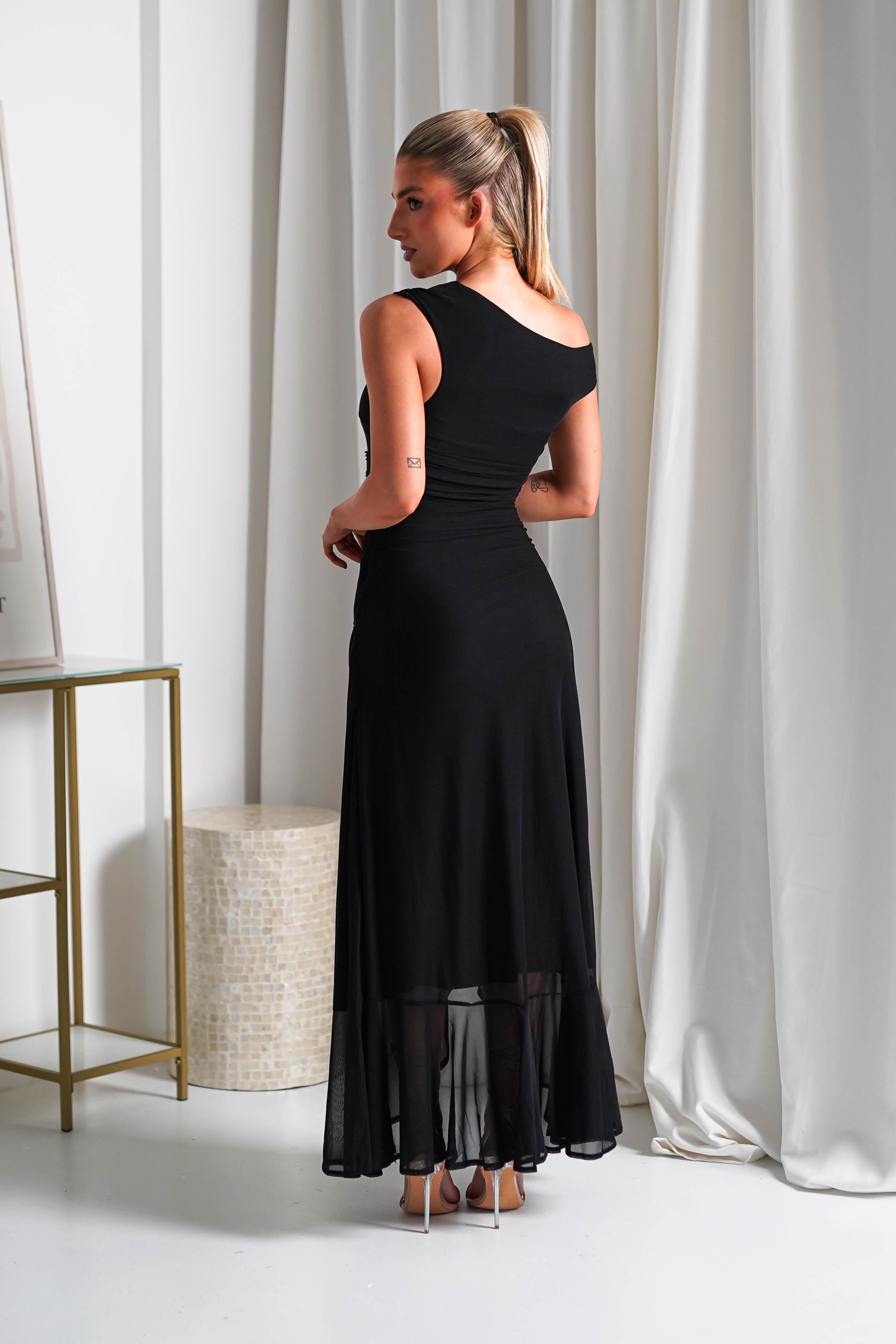 A woman in a form-fitting, one-shoulder midaxi dress stands in a room with white curtains. She is looking to the side, showing her profile. The dress features a sheer hem and stretchy mesh fabric. Her hair is pulled back in a ponytail. A table and abstract artwork are in the background.