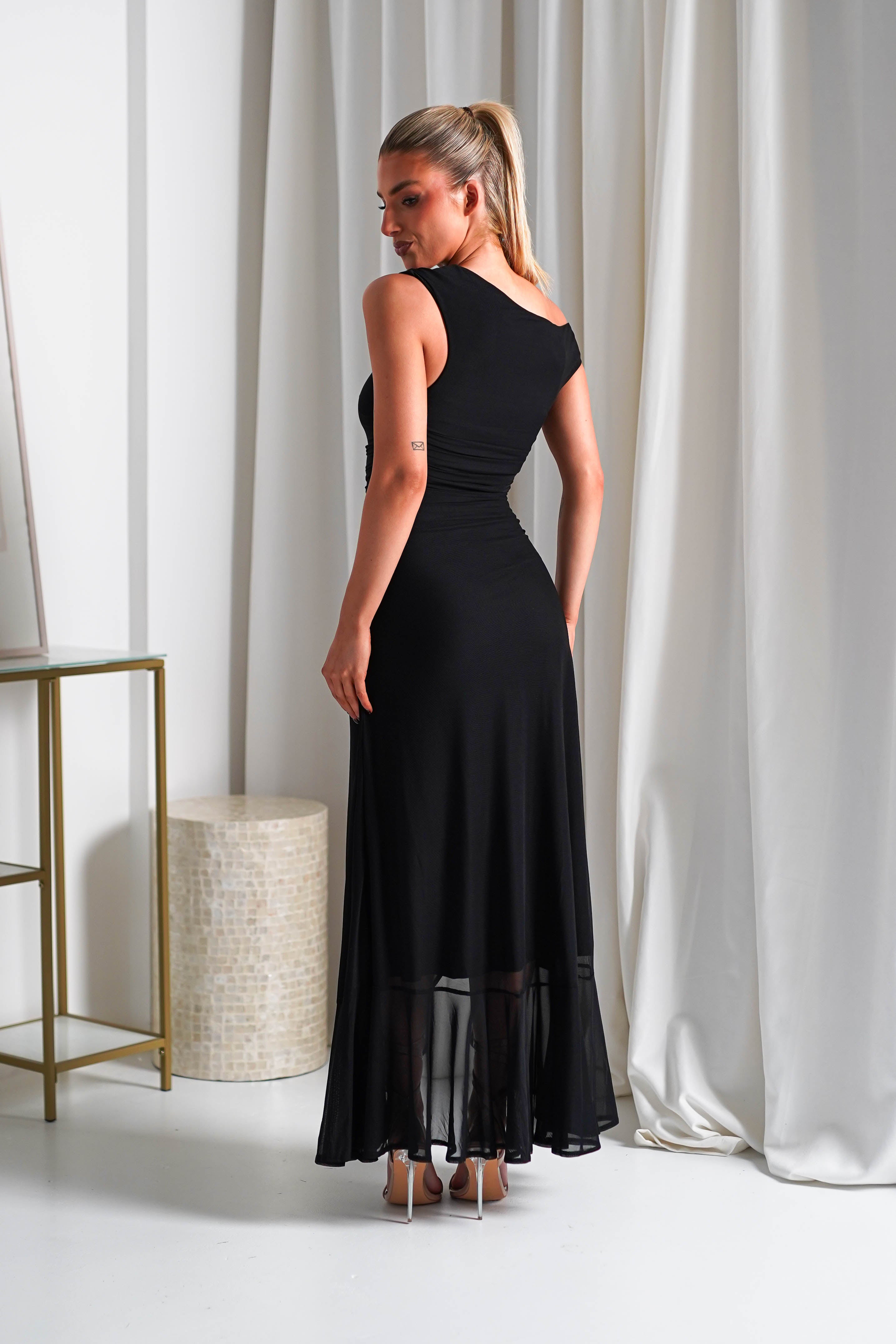 A person in a black sleeveless midaxi dress with a stretchy mesh fabric stands facing away, looking over their shoulder. The dress has a sheer hem and they are on high heels. The room features white curtains and a small table adorned with a textured cylindrical object.