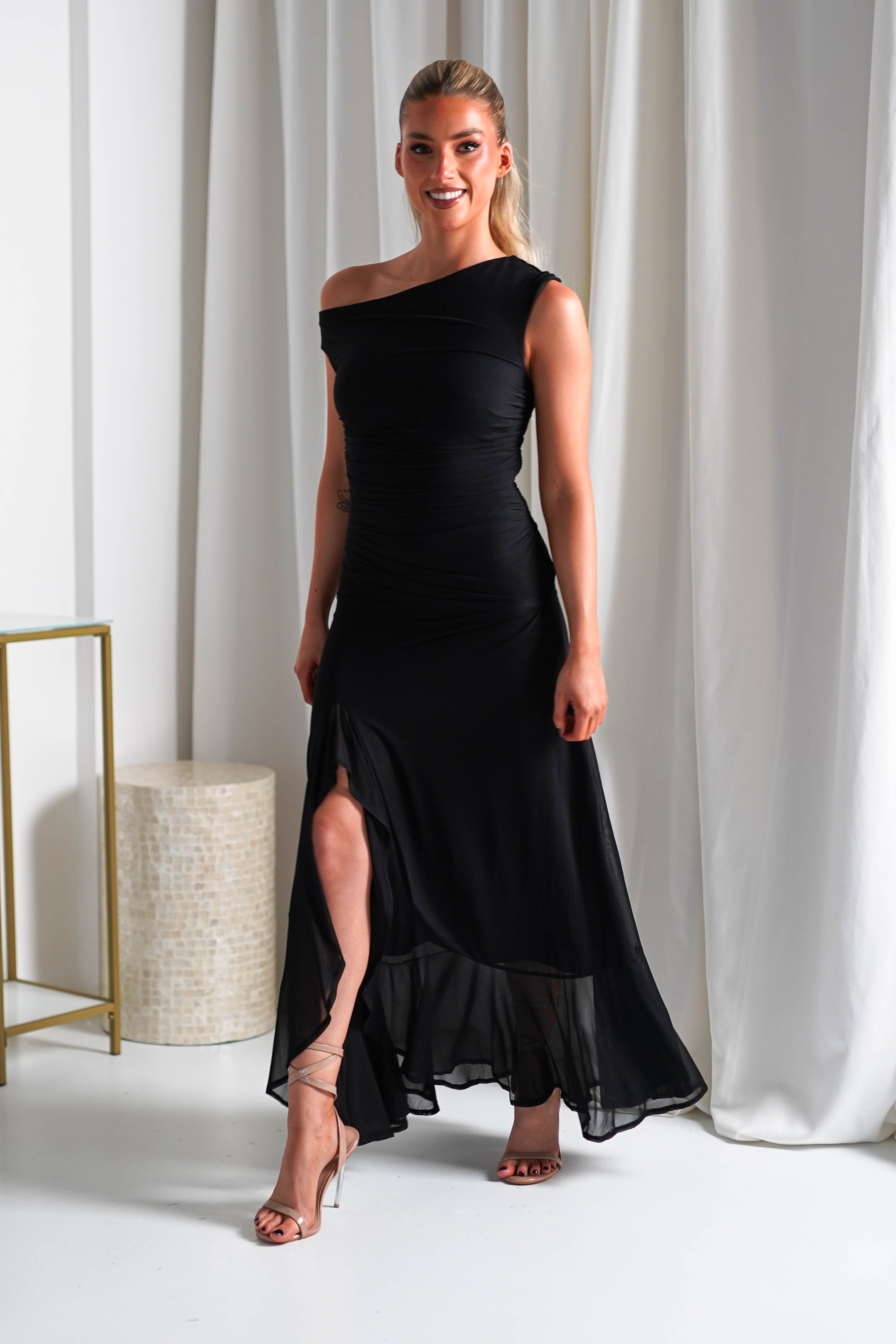Woman in an elegant, off-the-shoulder black midaxi dress with a side slit, standing in a bright room with white curtains and a small table. Ideal wedding guest attire, she pairs it with strappy heels and has her hair pulled back. The stretchy mesh fabric adds comfort to her chic look.