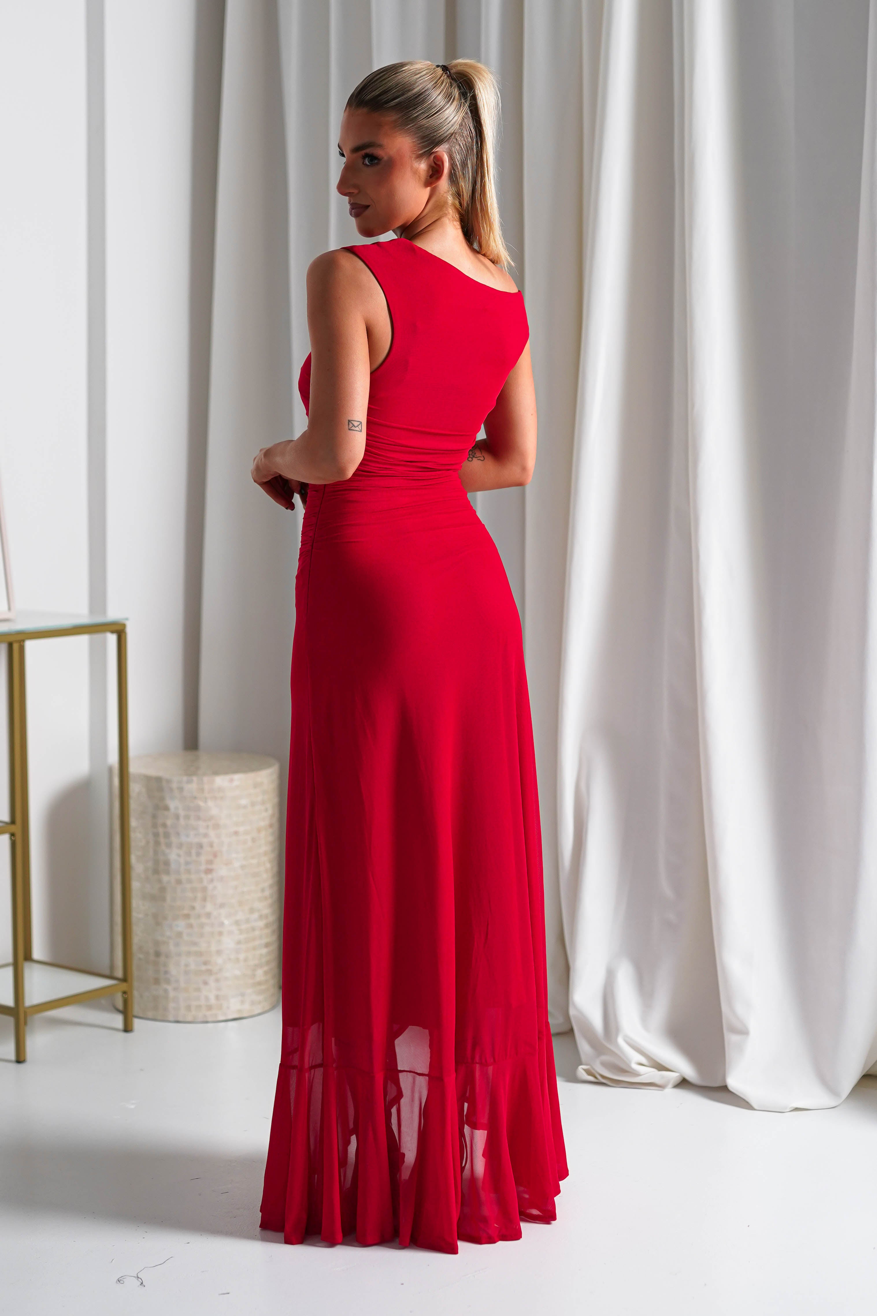 A woman with blonde hair in a ponytail wears a sleeveless, floor-length red midaxi dress crafted from stretchy mesh fabric, standing indoors with her back mostly to the camera, against white curtains and minimalist decor.