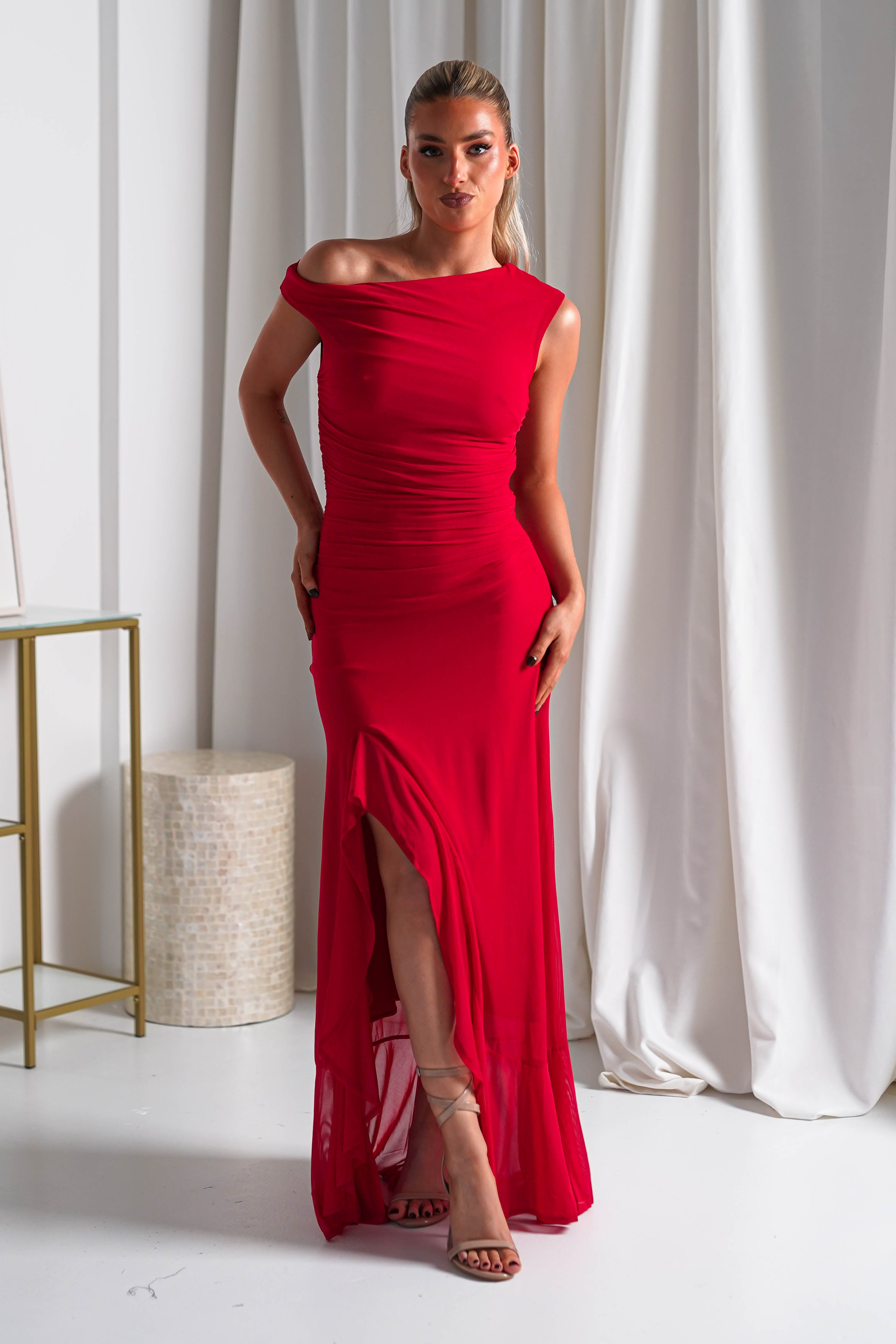 A woman in a fitted, off-the-shoulder red midaxi dress made from stretchy mesh fabric stands confidently indoors in strappy nude heels, with white curtains and minimal furniture in the background—perfect as wedding guest attire.