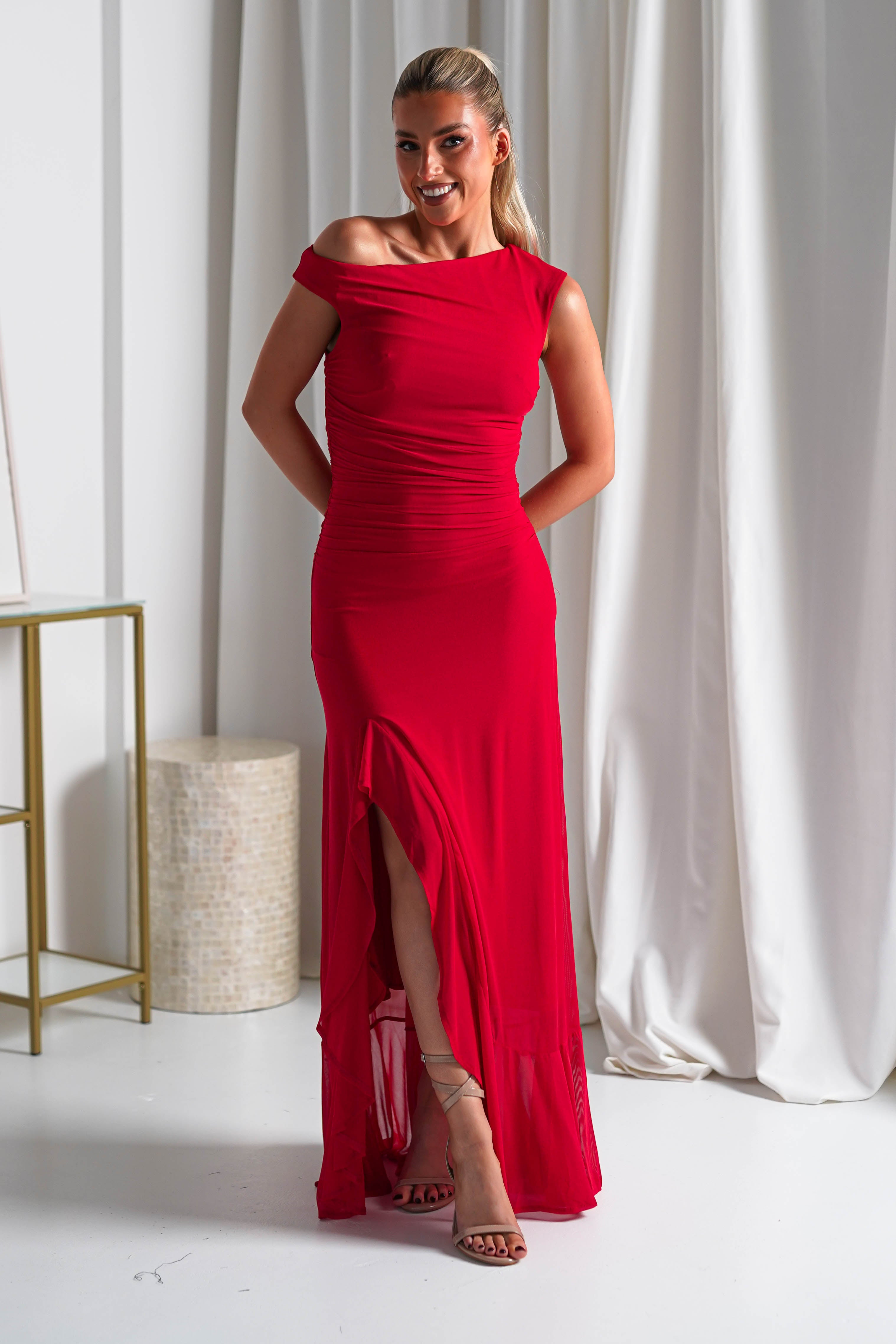 A woman smiles while wearing a fitted, off-the-shoulder red midaxi dress with a thigh-high slit. Perfect as wedding guest attire, she stands indoors on a white floor in front of white curtains, with her hands behind her back.