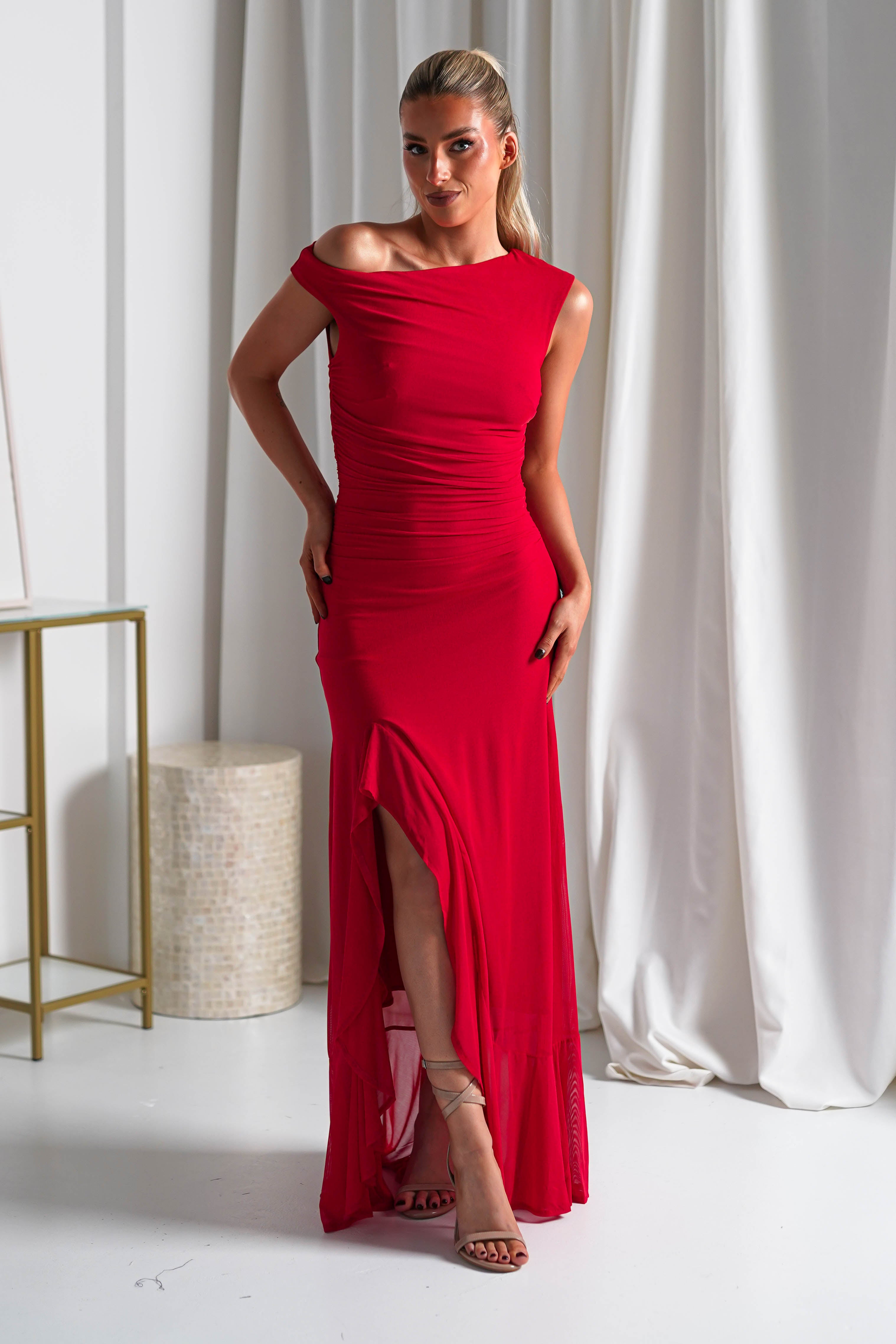 A woman stands indoors wearing a fitted, off-the-shoulder red midaxi dress with a thigh-high slit. She has long, blonde hair pulled back and poses with her hands on her hips in front of white curtains—perfect wedding guest attire.