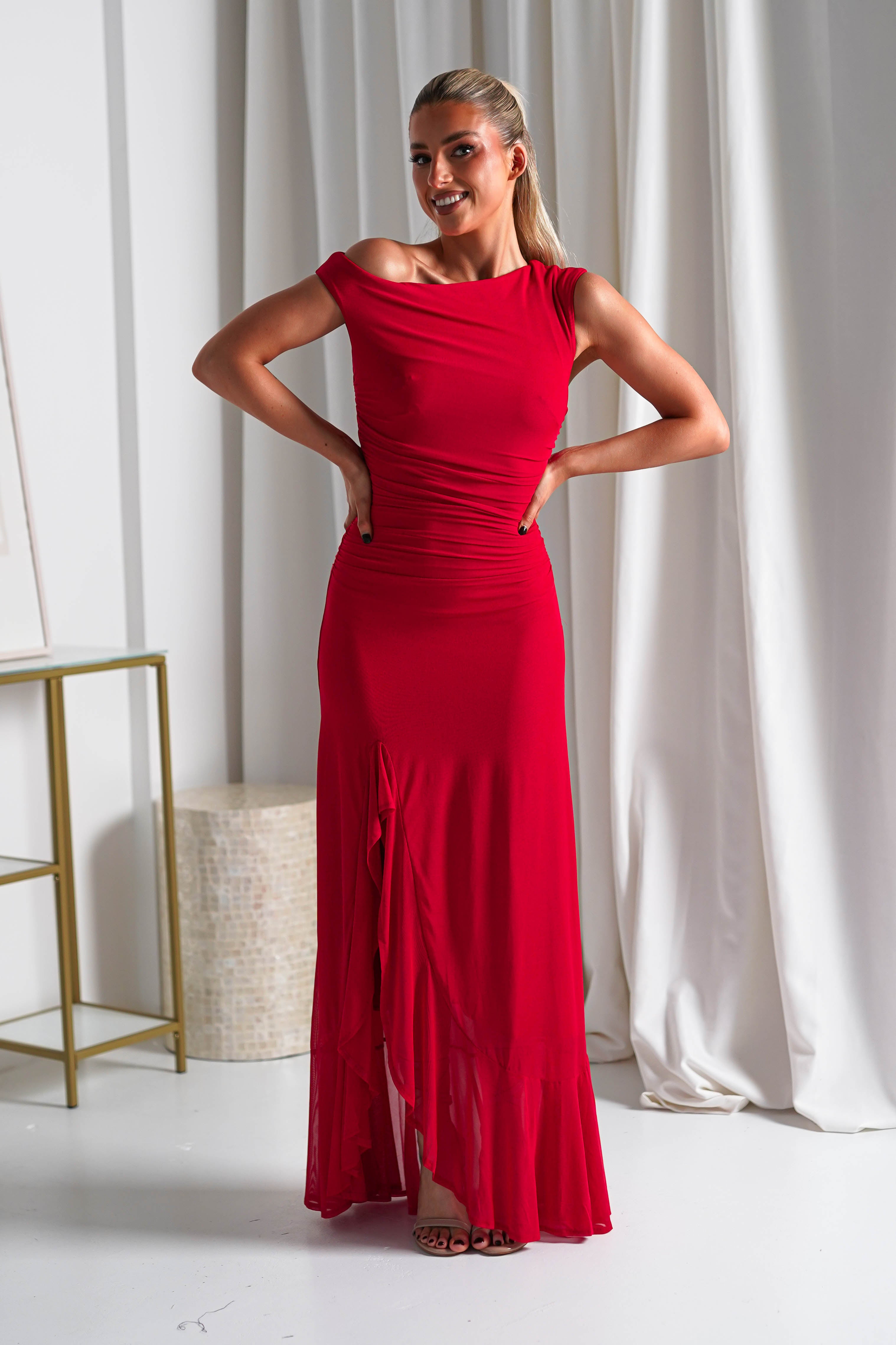 A woman in a fitted, off-the-shoulder midaxi dress stands indoors with hands on her hips, smiling. White curtains and modern furniture provide a chic backdrop, making this look ideal for wedding guest attire.