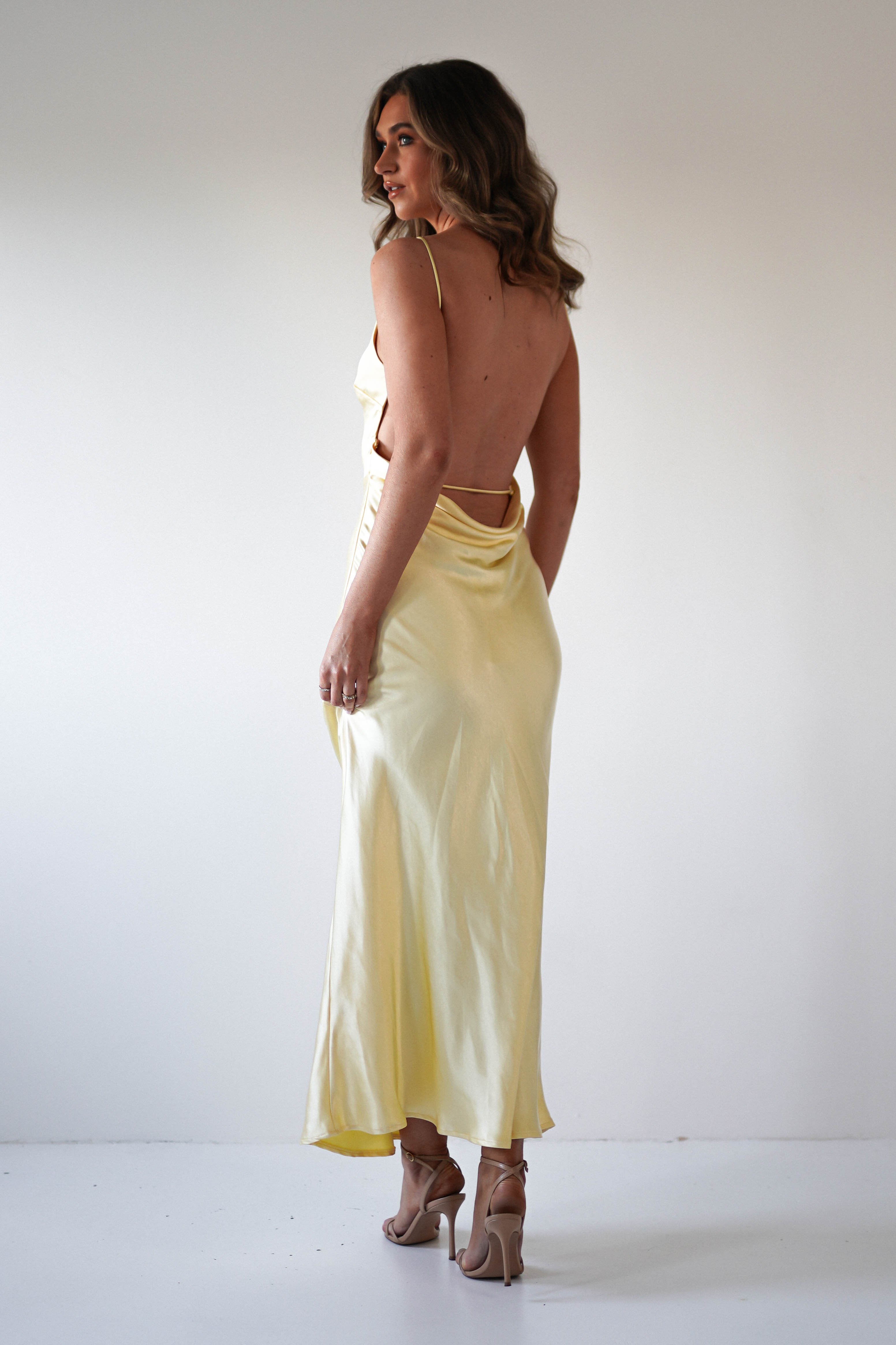 A woman with wavy hair wears a light yellow, strapless satin maxi dress and nude high heels, standing on a white floor and facing away from the camera against a plain white background—perfect for wedding guest or special occasions.