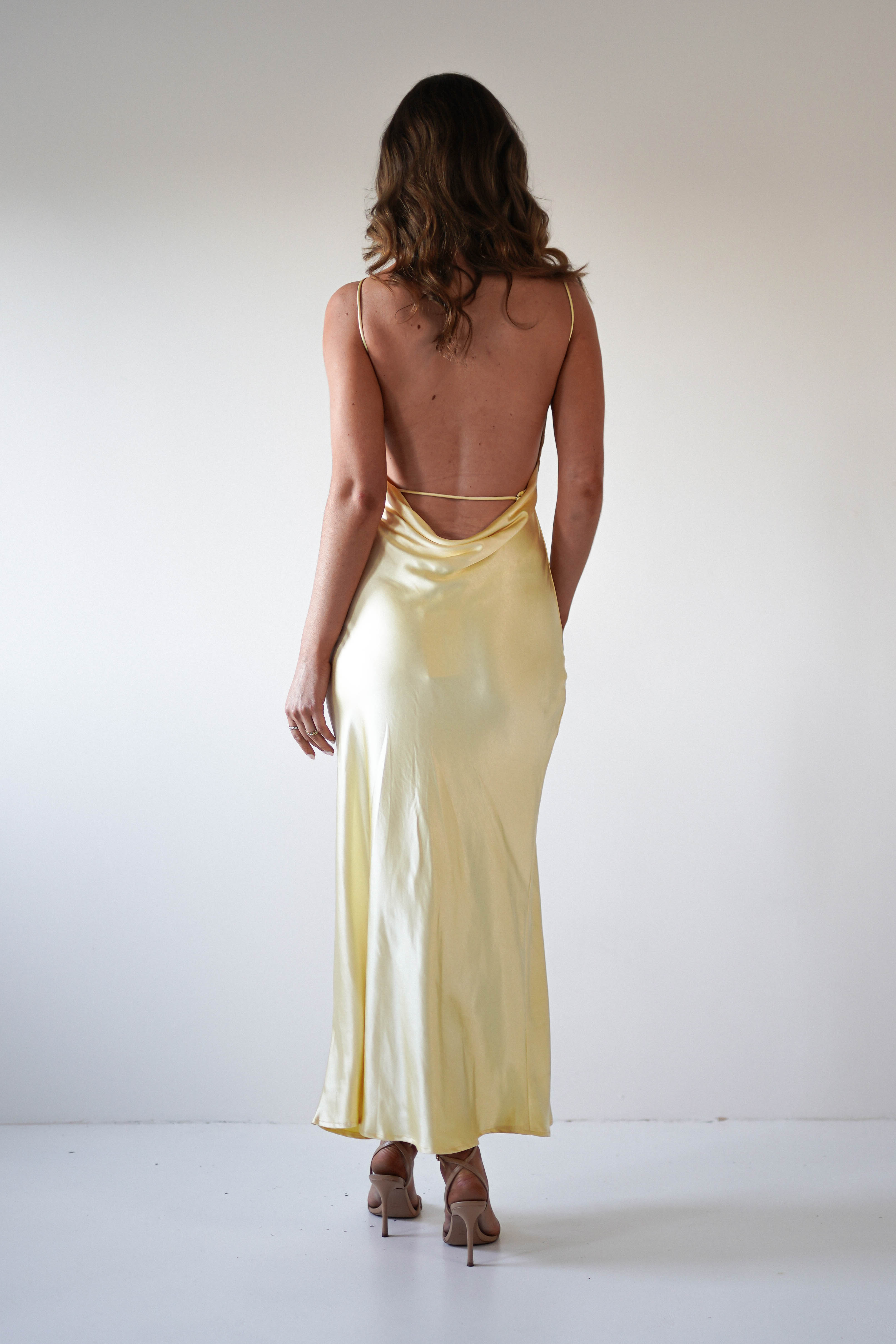 A woman in a backless, pale yellow strapless satin maxi dress and beige heels stands facing away from the camera against a plain white background—an elegant look perfect for wedding guest attire or special occasions.