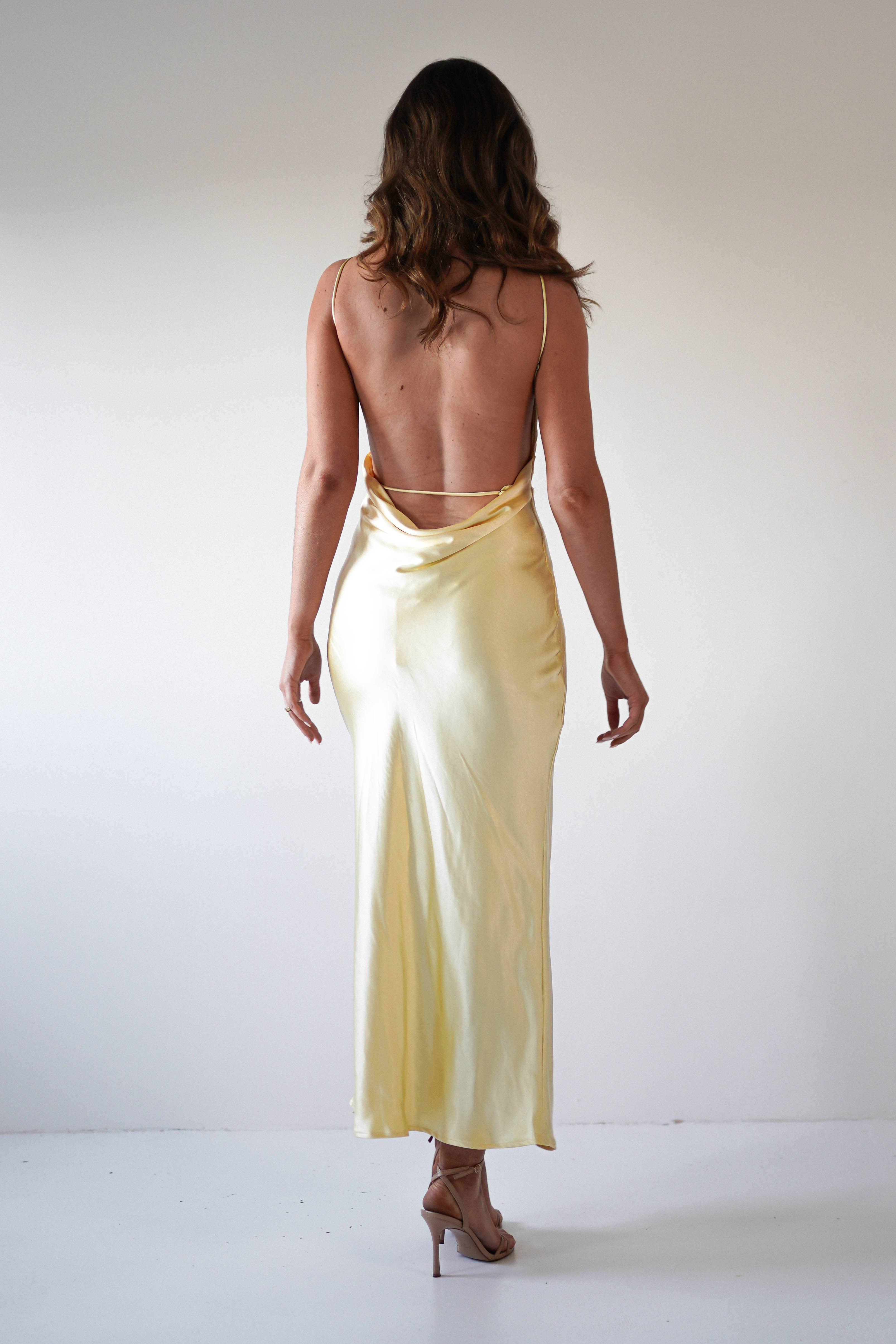 A woman with wavy brown hair stands facing away, wearing a long, strapless satin maxi dress in pale yellow and beige high-heeled sandals—an elegant look perfect for special occasions or as a stunning wedding guest ensemble.