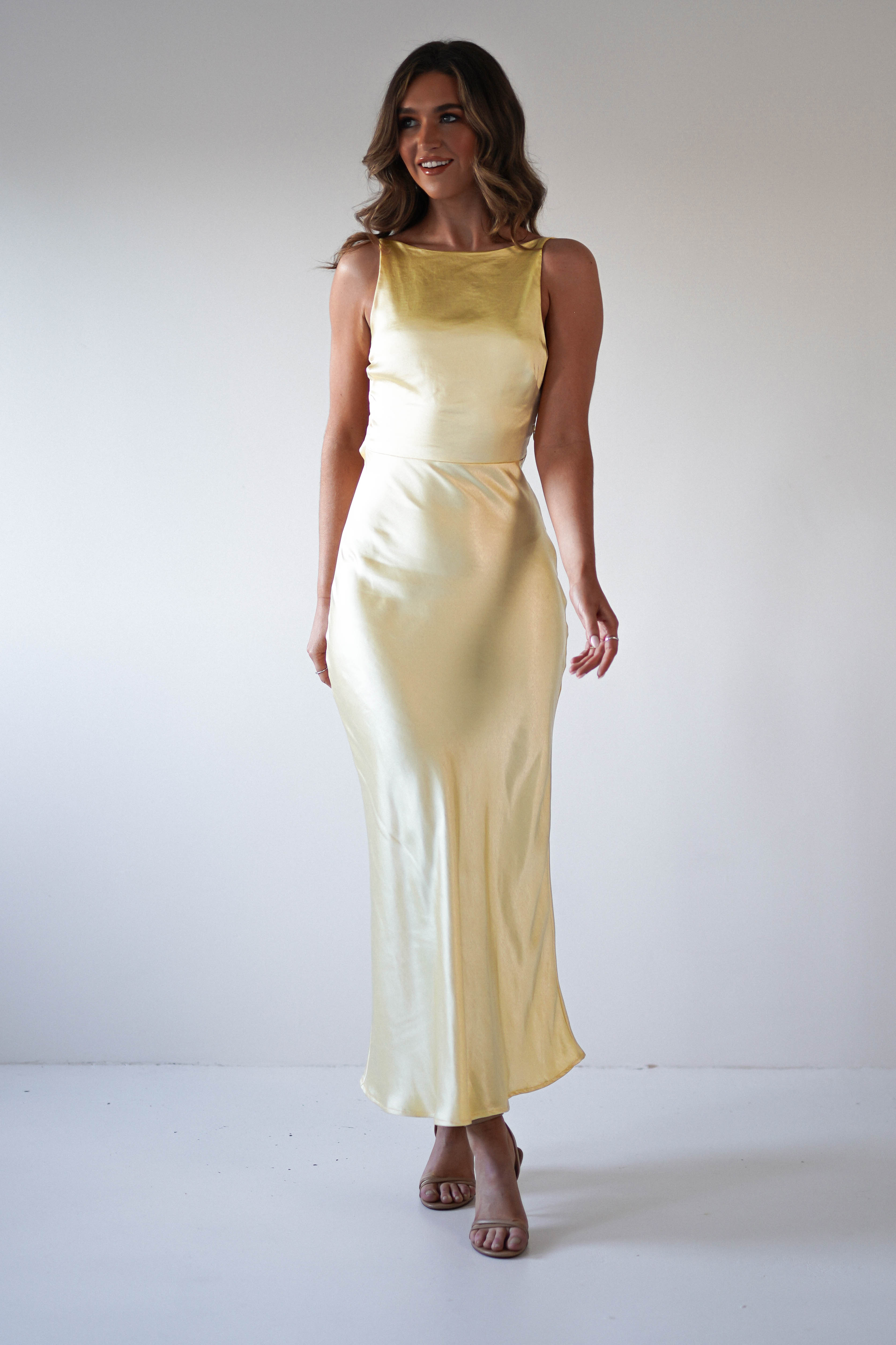A woman stands against a plain white background, smiling in a strapless satin maxi dress in light yellow with nude heeled sandals. Her wavy brown hair is styled loosely—an elegant look perfect for special occasions or as a wedding guest.
