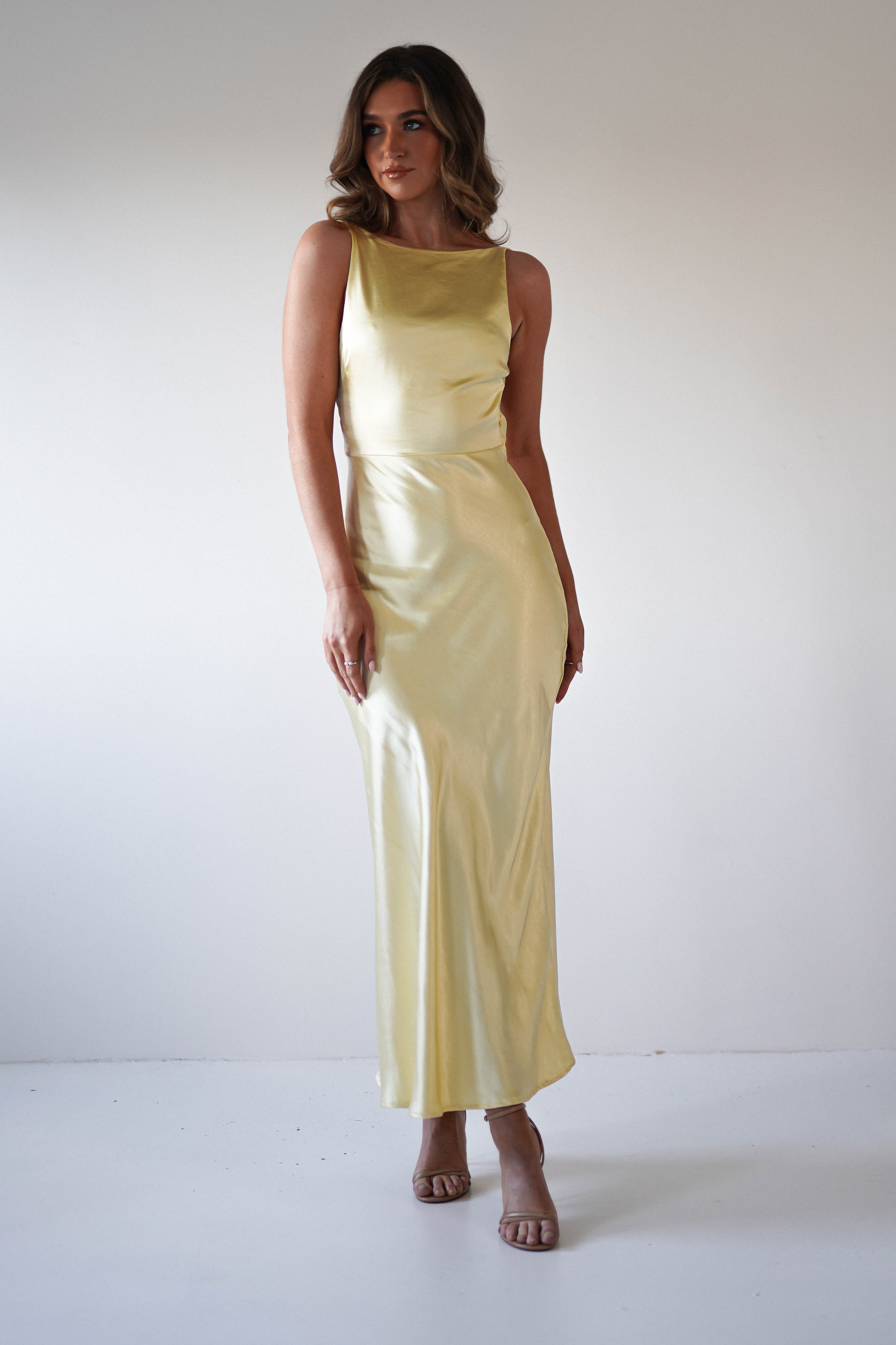 A woman stands in a minimalist setting wearing a strapless satin maxi dress in pale yellow, perfect for wedding guest attire or special occasions. She has wavy hair and open-toe heels, with the dress’s fitted waist adding elegant flair.
