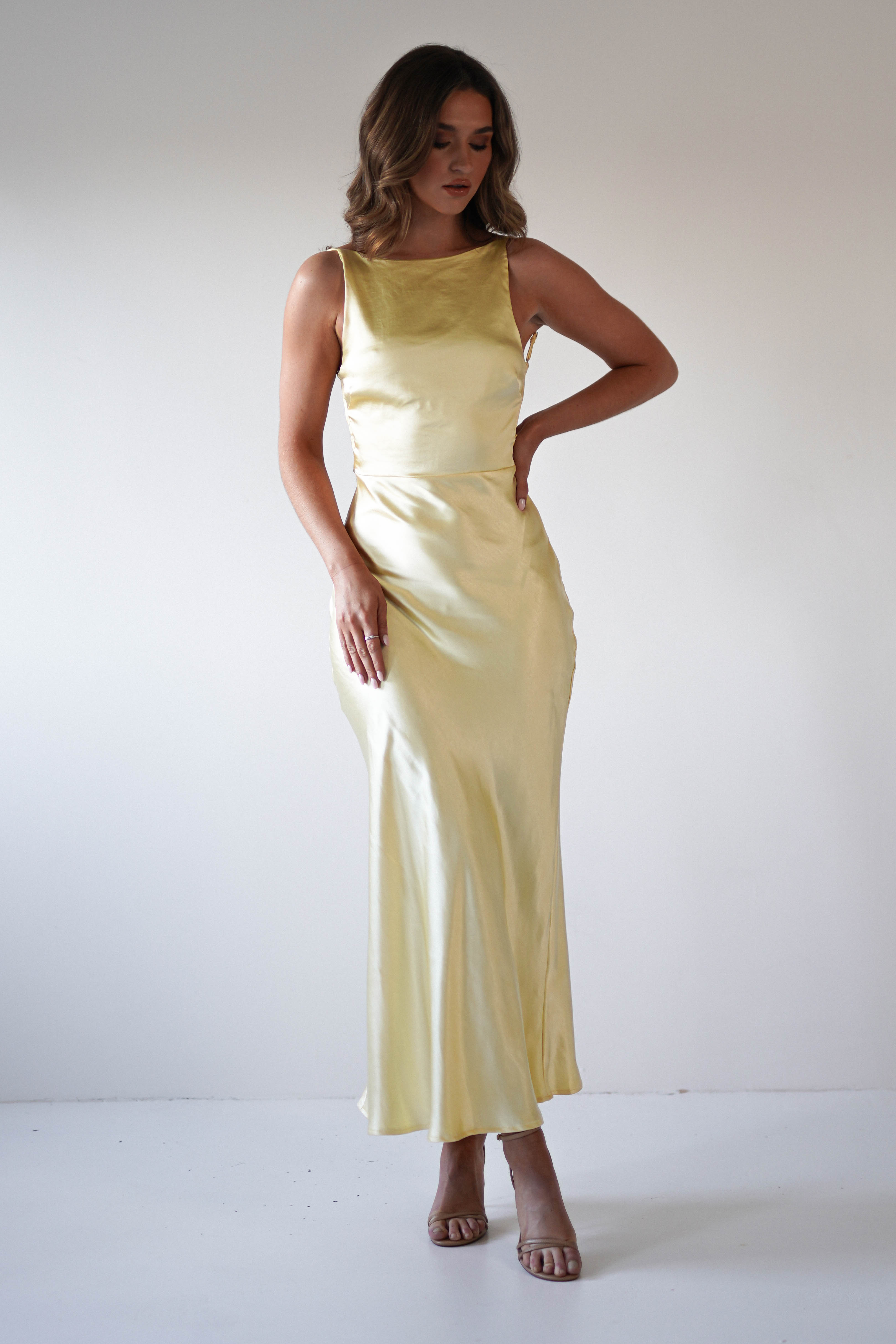 A woman stands against a plain white background, wearing a strapless satin maxi dress in pale yellow—perfect for special occasions or as a stunning wedding guest look. She pairs it with beige heeled sandals and has wavy, shoulder-length hair.