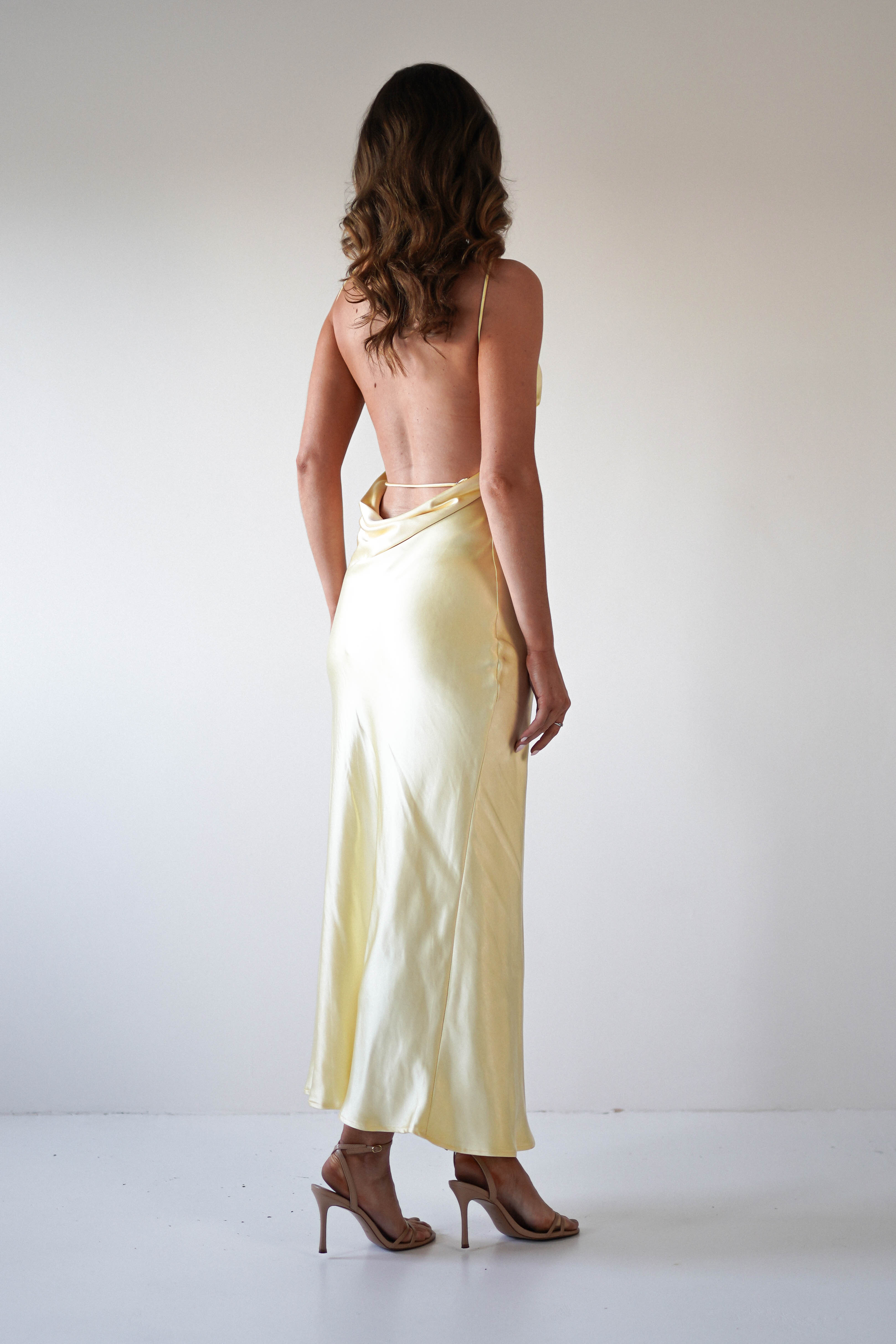 A woman with wavy brown hair wears a strapless satin maxi dress in light yellow and tan high heels, standing in a minimal white room facing away from the camera—the perfect look for special occasions or as a wedding guest.