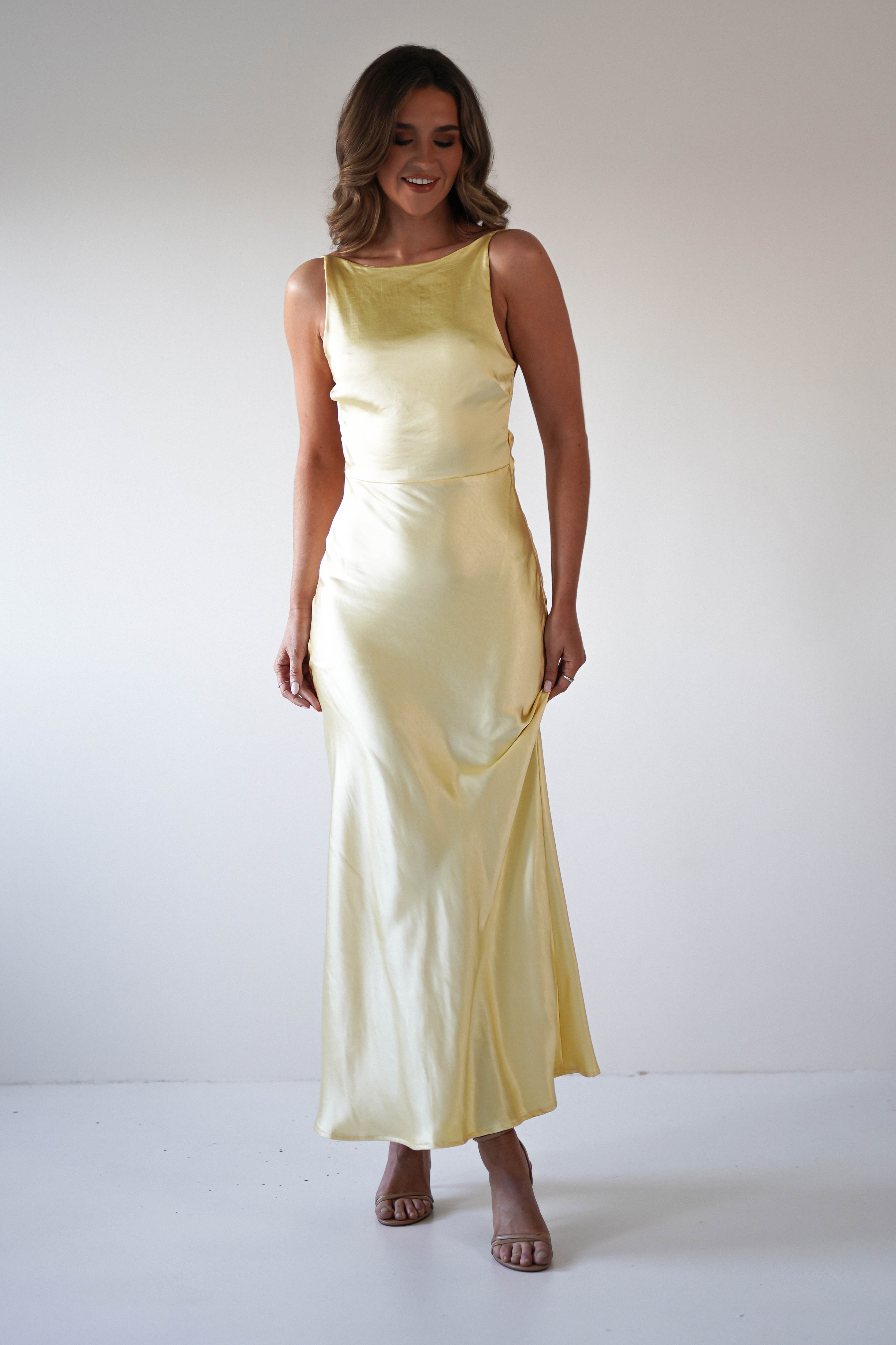 A woman in a strapless satin maxi dress stands smiling against a plain light background. Perfect for special occasions or as a wedding guest look, she pairs her outfit with wavy brown hair and open-toed, nude-colored heeled sandals.