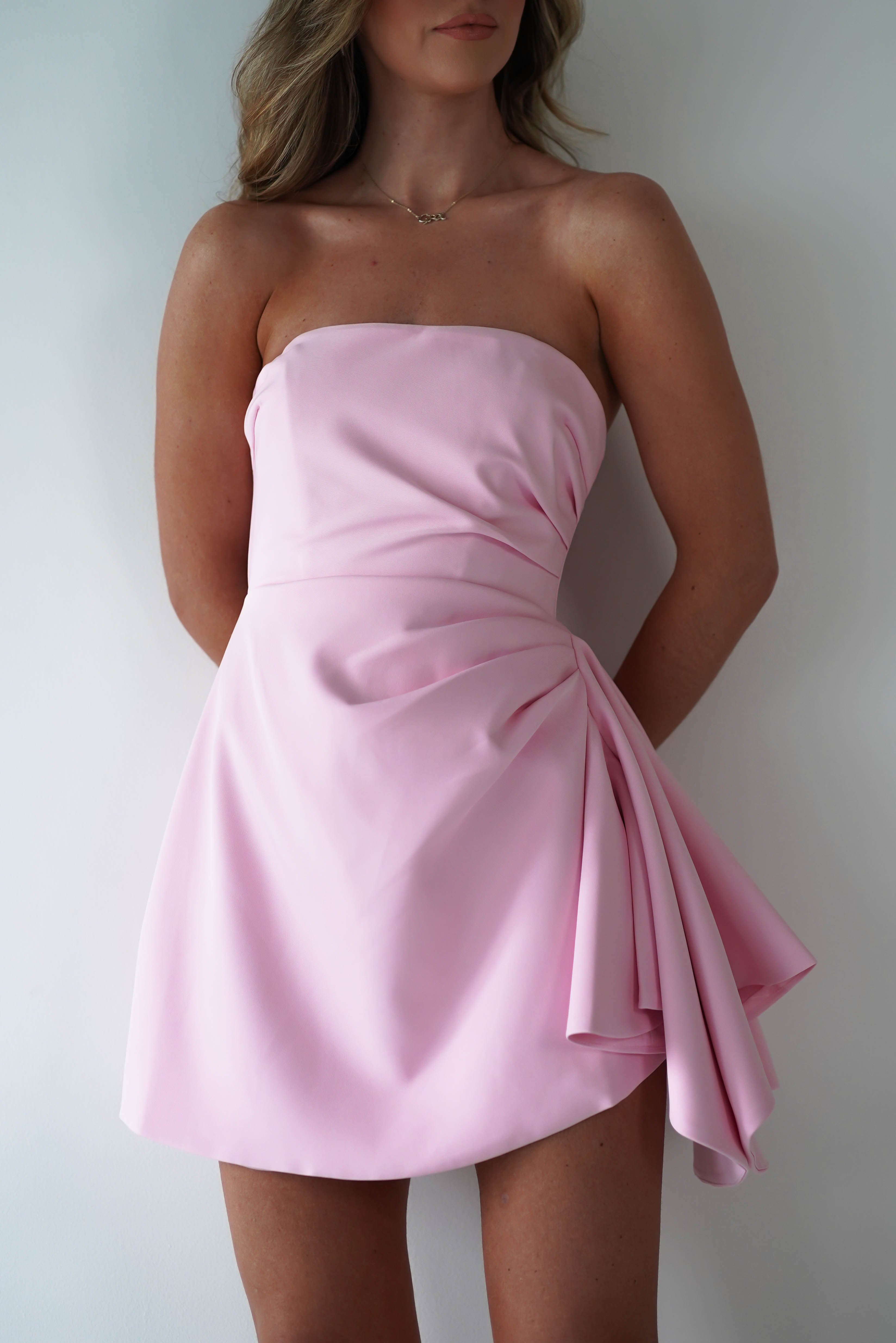 A woman wearing a strapless mini dress in light pink satin with draped detailing on one side, stands against a plain white background. Her face is partially out of frame, showcasing this chic party dress perfect for any celebration.