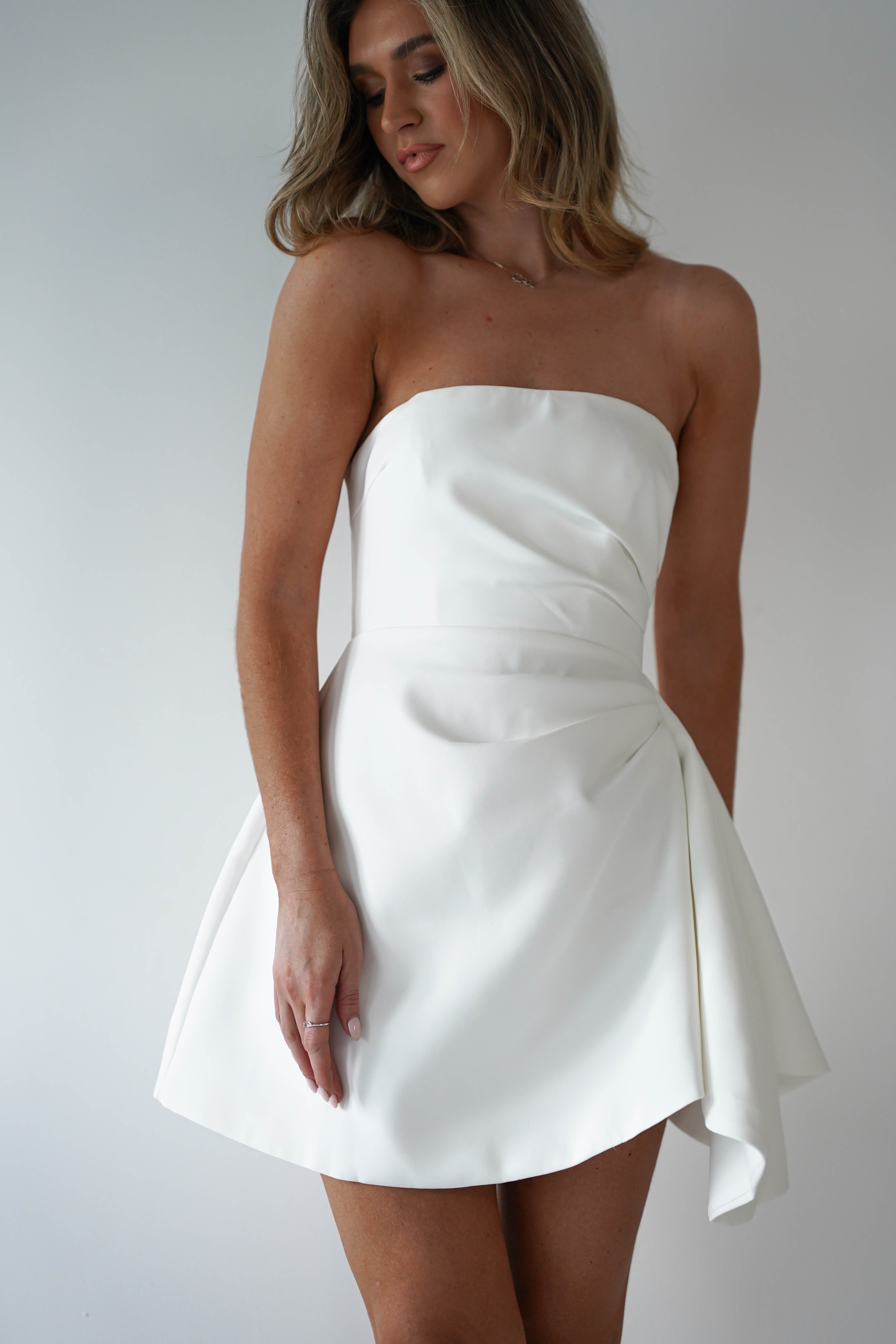 A person wearing a strapless, white, asymmetrical mini dress poses against a plain background. The party dress features a fitted bodice and flares out at the skirt. The person has shoulder-length hair and stands slightly turned to the side.