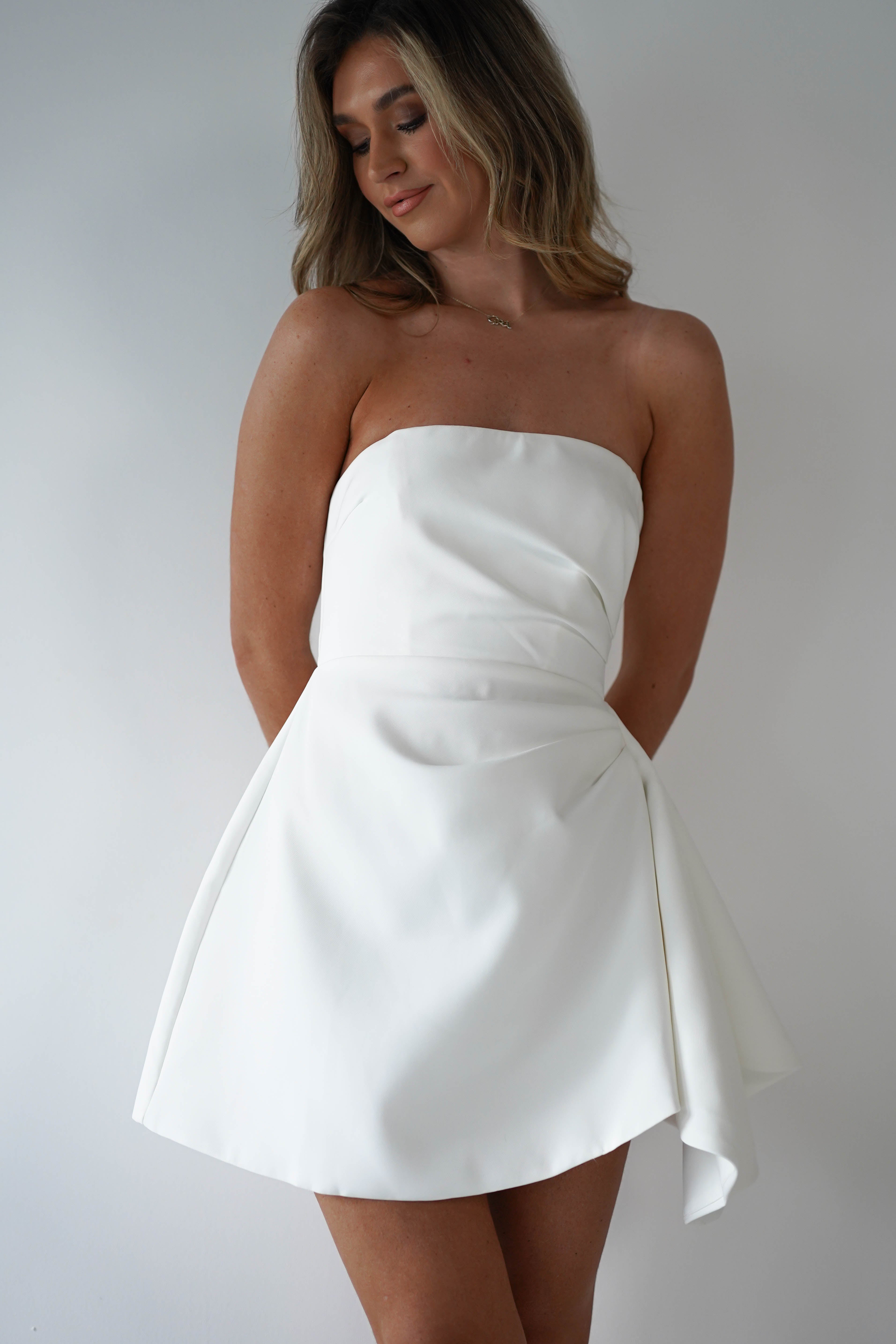 A person wearing a strapless mini dress in white stands against a plain background, looking down. The polyester dress features a fitted bodice and elegantly flares out from the waist, exuding understated charm.