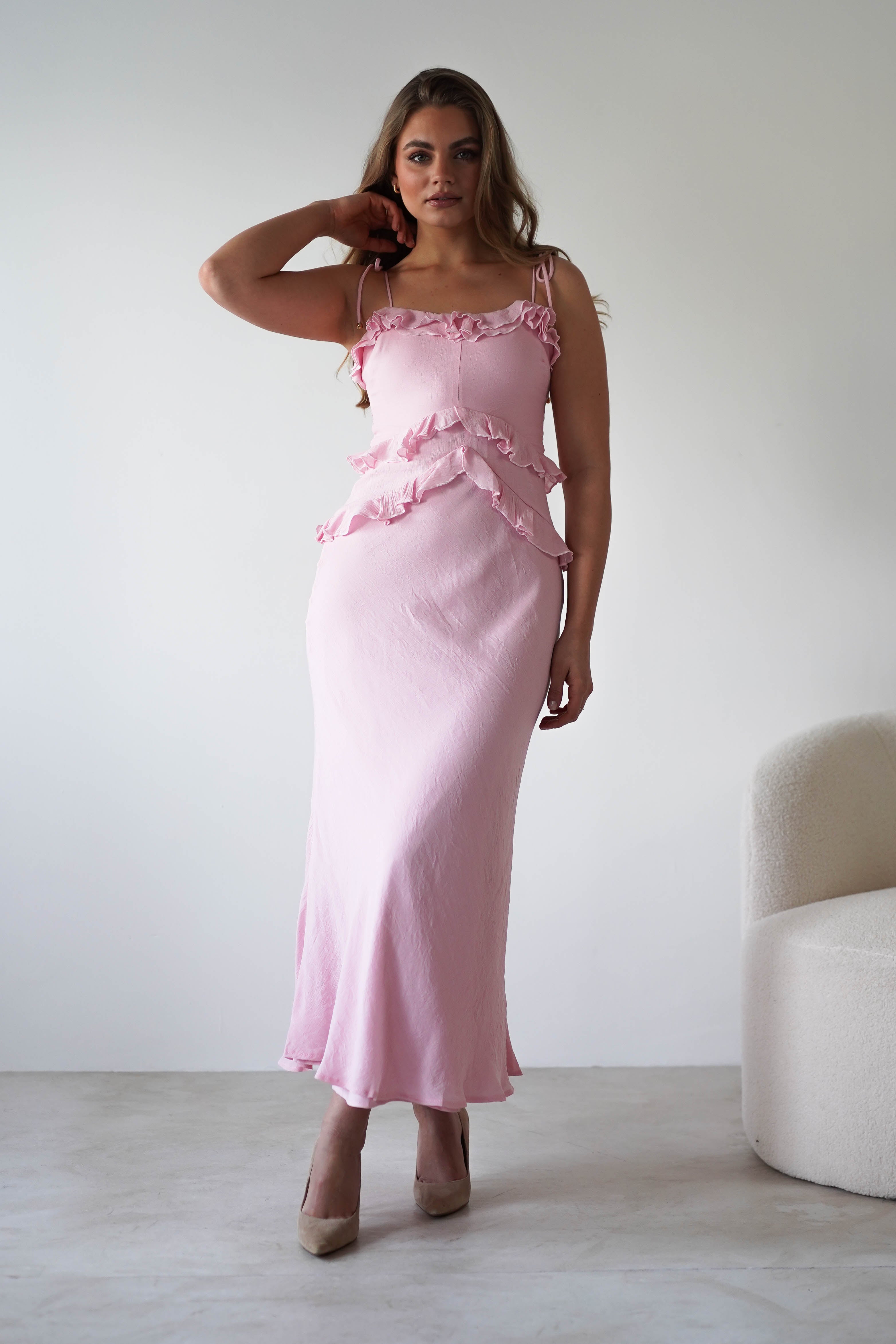 A woman wearing a light pink ruffled maxi dress with adjustable tie straps stands indoors on a light floor beside a white textured chair, posing with one hand touching her hair against a plain, light background. Perfect as a wedding guest dress.