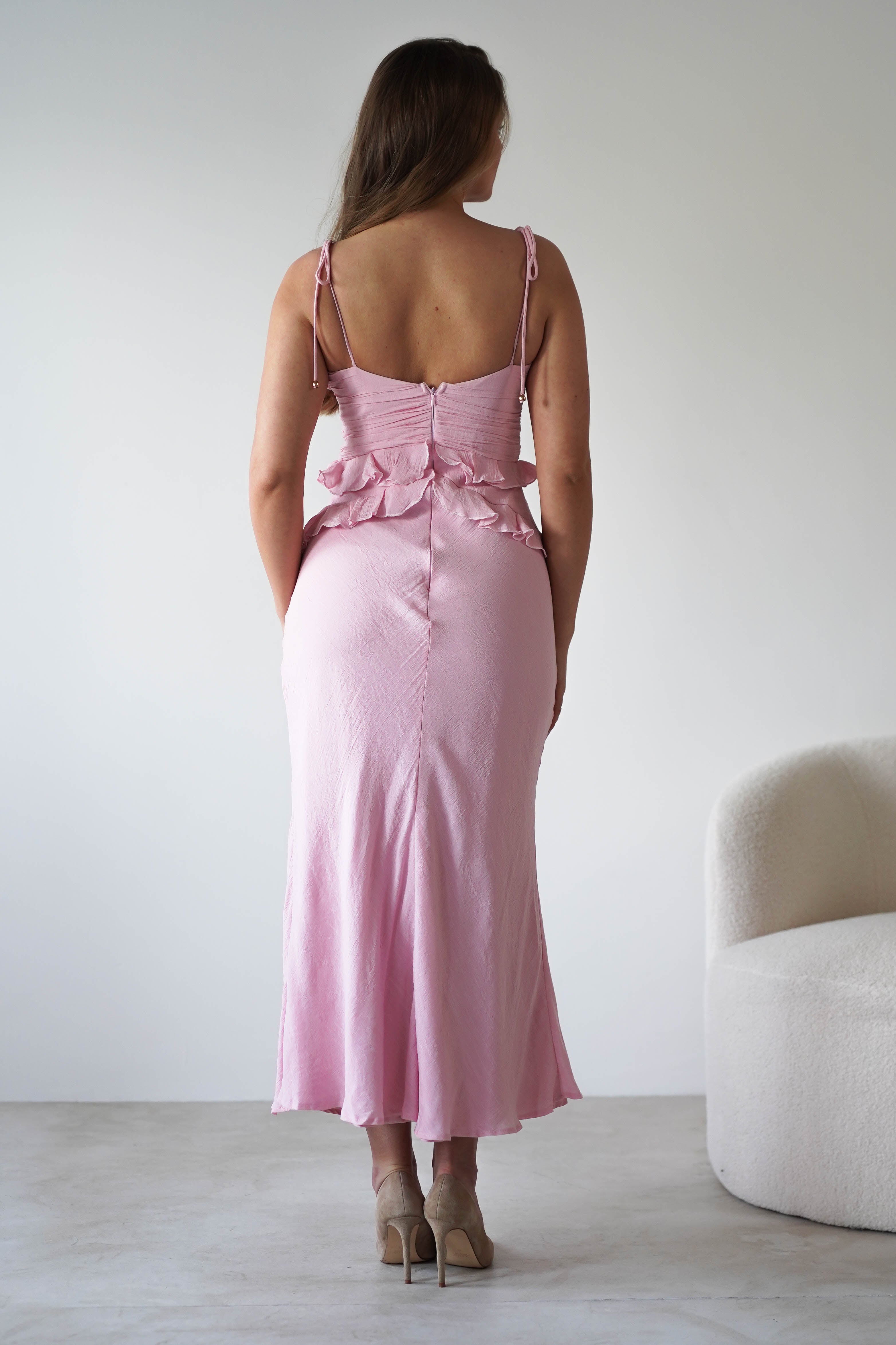 A woman stands facing away, wearing a light pink maxi dress with ruffle details at the waist and adjustable tie straps. She is on a light floor near a white, textured armchair against a plain wall—perfect as a wedding guest dress.