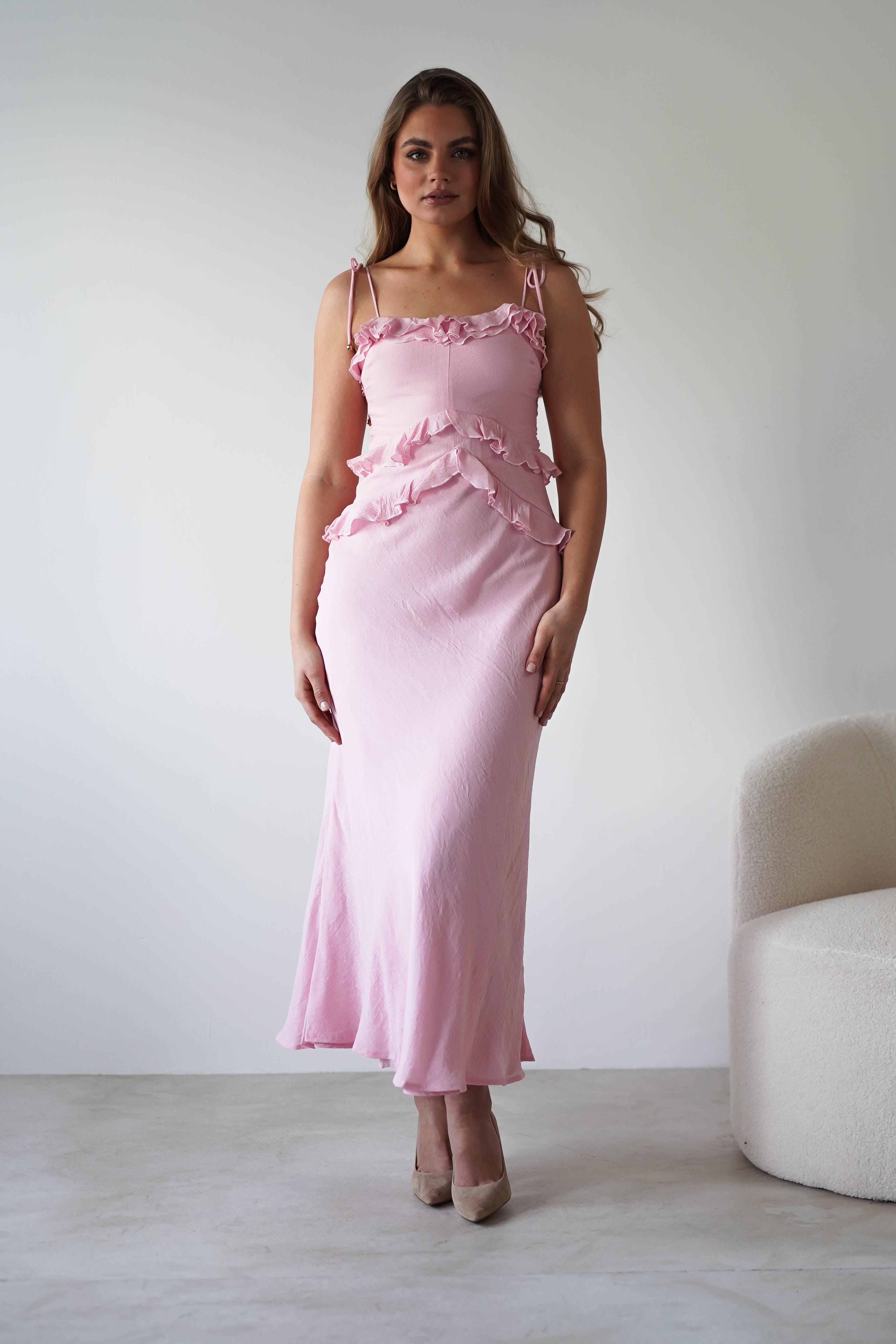 A woman stands indoors wearing a fitted, light pink wedding guest dress with ruffle details and beige heels. The sleeveless style features adjustable tie straps. She is near a white curved chair against a plain white wall.