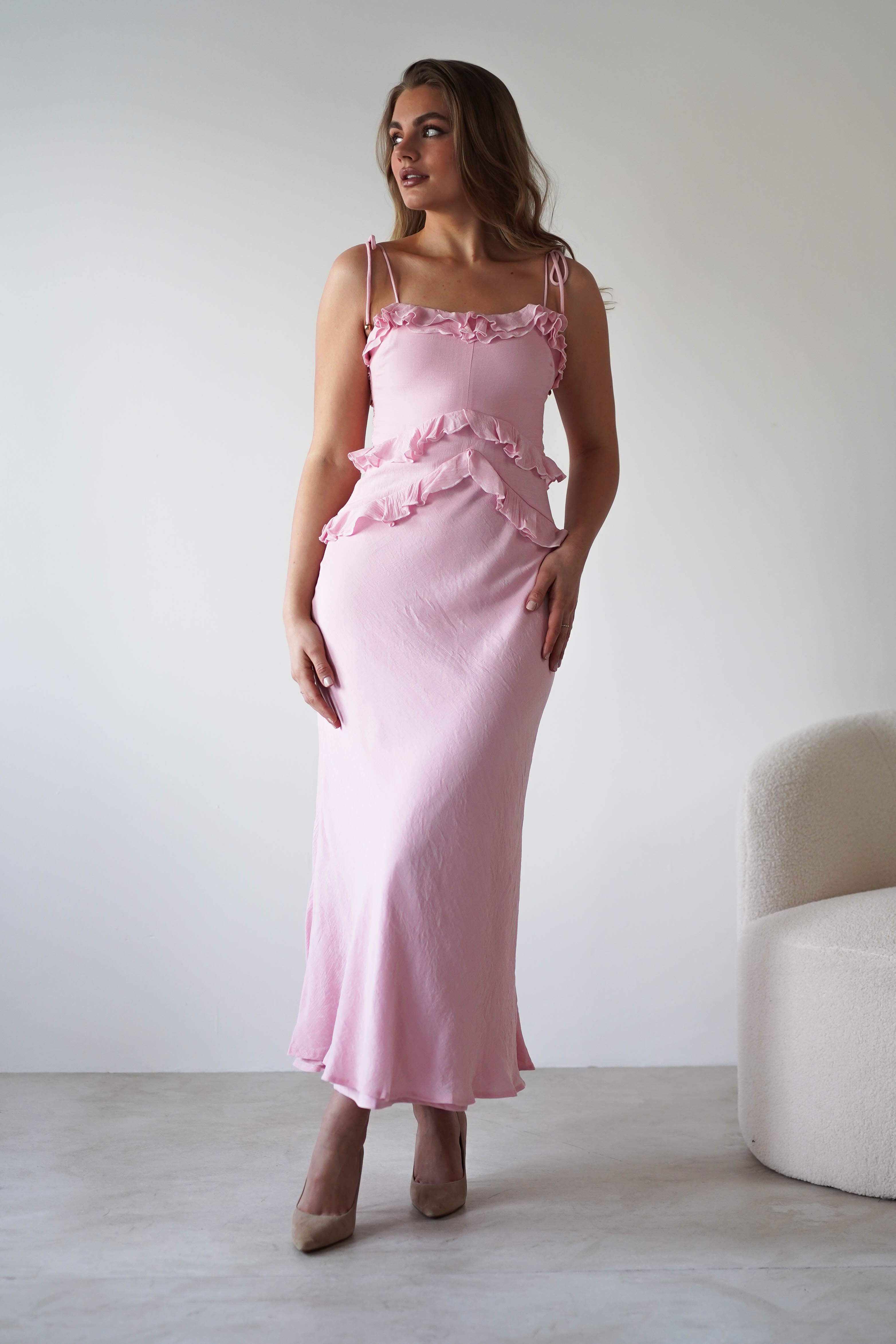 A woman wearing a long, light pink ruffled maxi dress with adjustable tie straps and beige heels stands indoors near a cream-colored chair, posing against a plain light background. Perfect as a chic wedding guest dress.