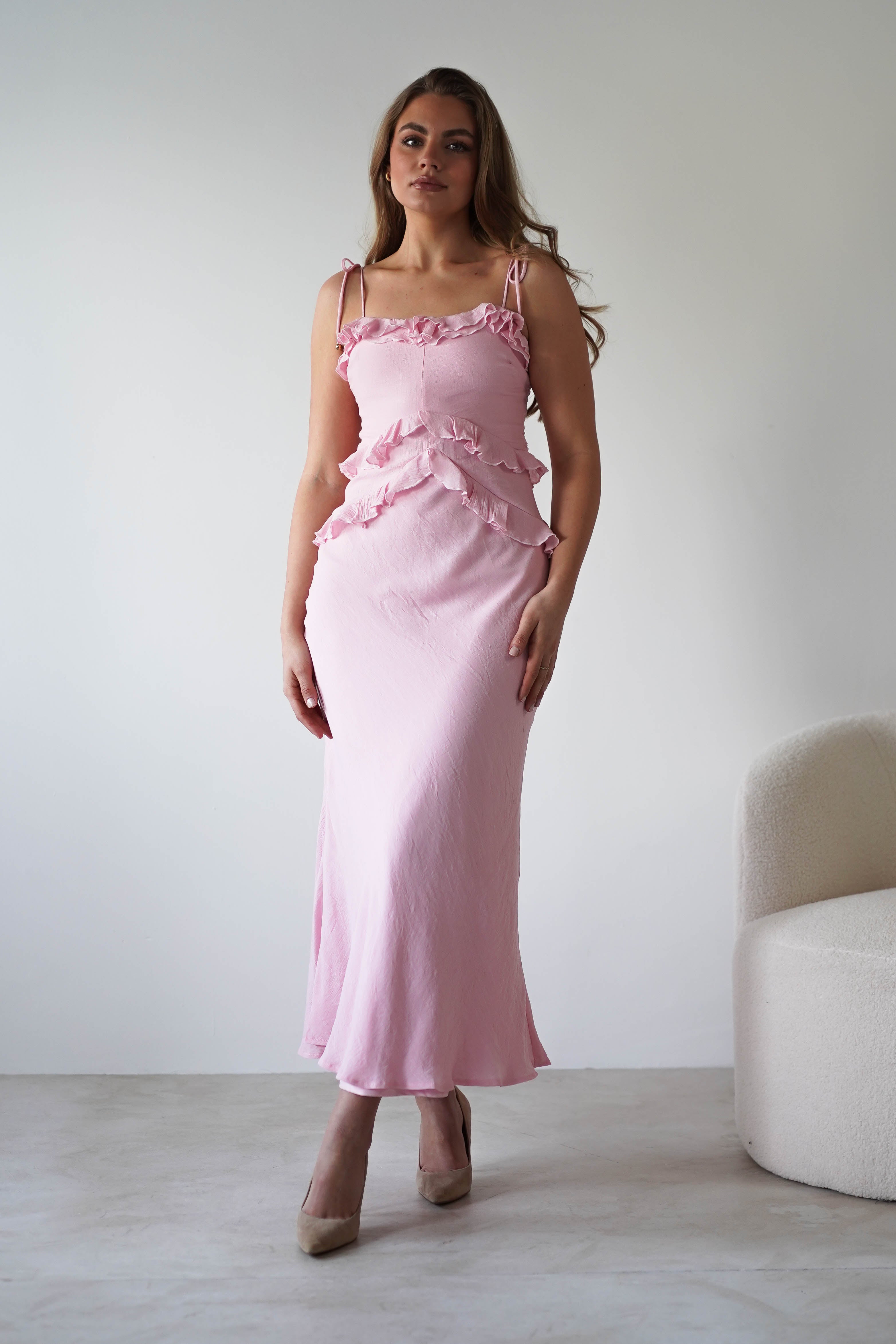 A woman stands indoors wearing a long, light pink maxi dress with ruffles and adjustable tie straps, paired with beige high heels. She is next to a cream-colored, curved chair against a plain, light-colored wall.