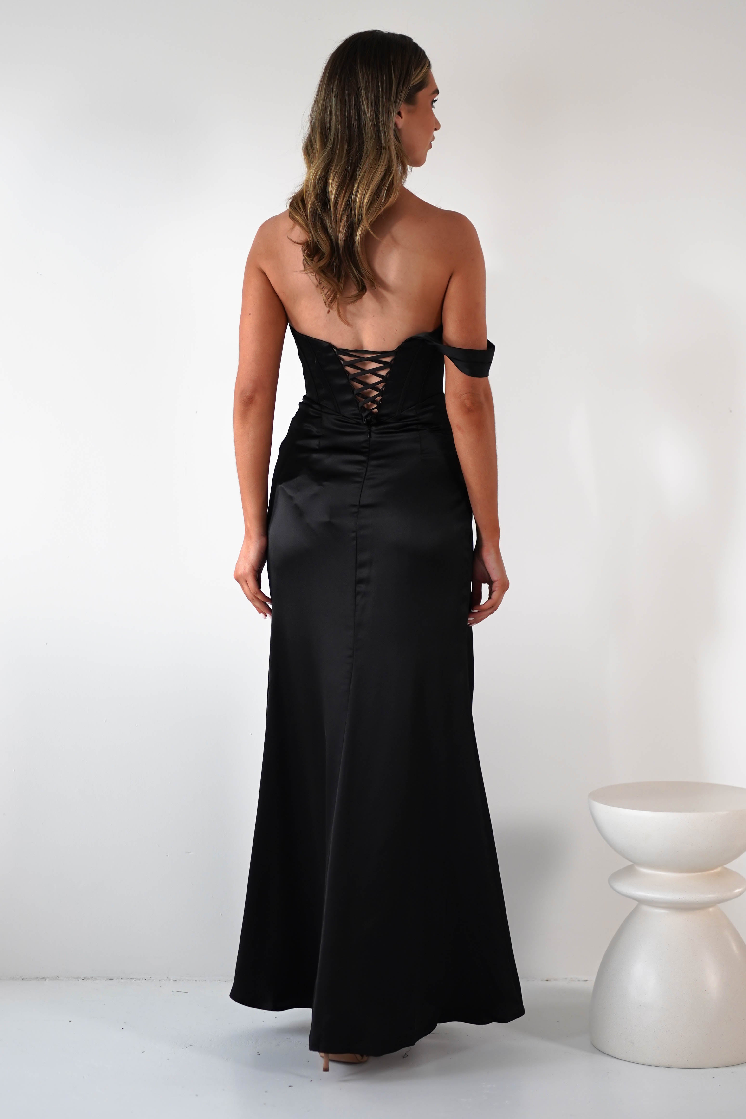 A woman stands facing away, wearing a long, strapless black corset dress with lace-up detailing on the back. She has wavy, shoulder-length hair and stands near a small, modern white stool against a plain white background.