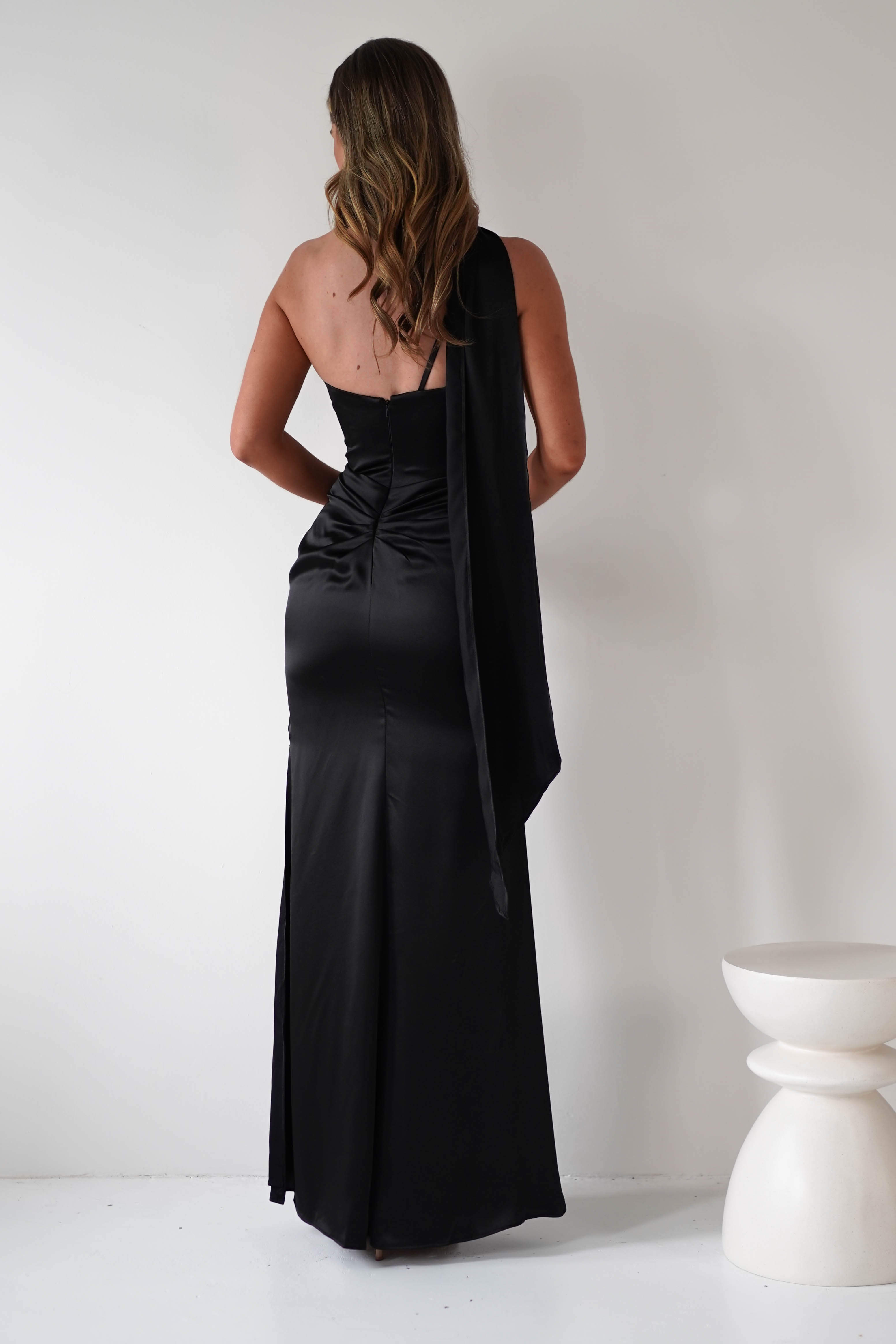 A woman with wavy hair stands facing a white wall, wearing a long, fitted black satin dress with an asymmetrical shoulder strap, gathered midriff, and draped fabric detail. A modern white stool is beside her.