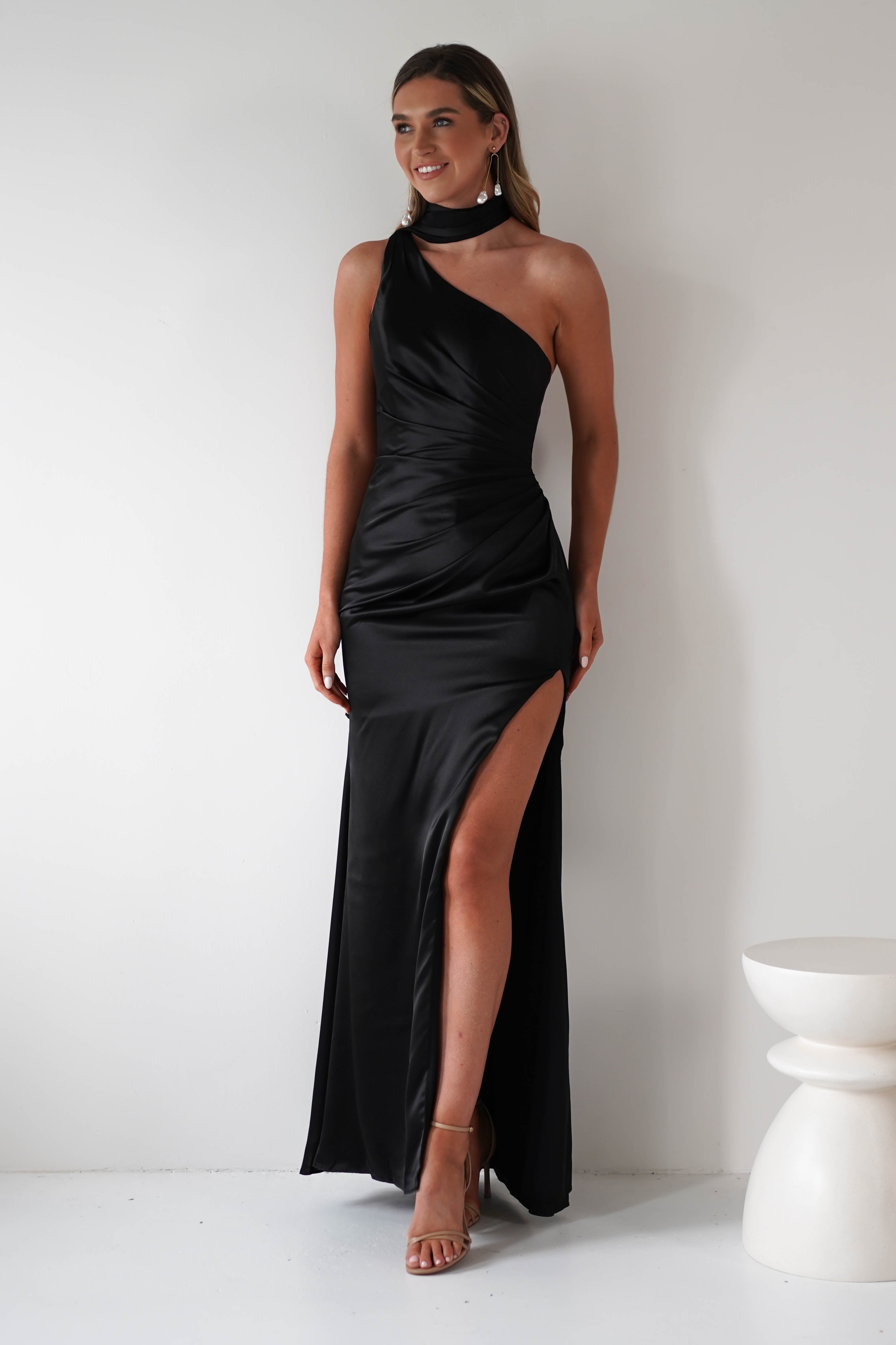 A woman poses in a sleek, black, one-shoulder satin gown with a gathered midriff and high thigh slit. She wears hoop earrings and nude heels, standing against a plain white wall next to a small white stool.